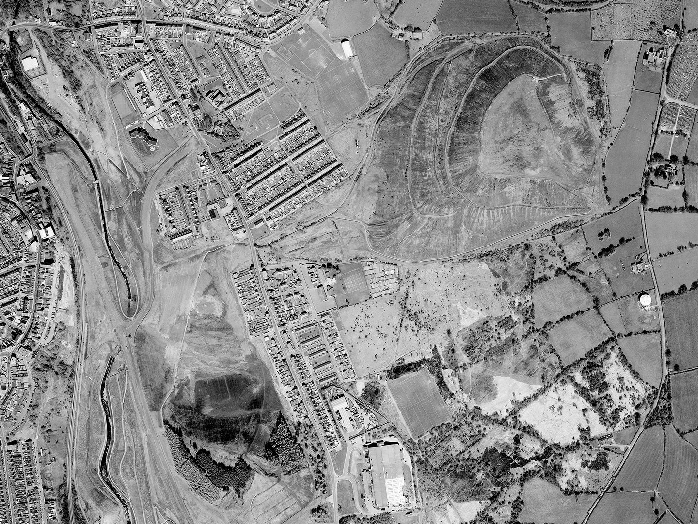 A historic black and white vertical aerial photo of a post-industrial landscape. A river runs along the left-hand edge of the image. A mix of housing and fields fills most of the image, along with two large coal spoil heaps at the top right and bottom left corners.