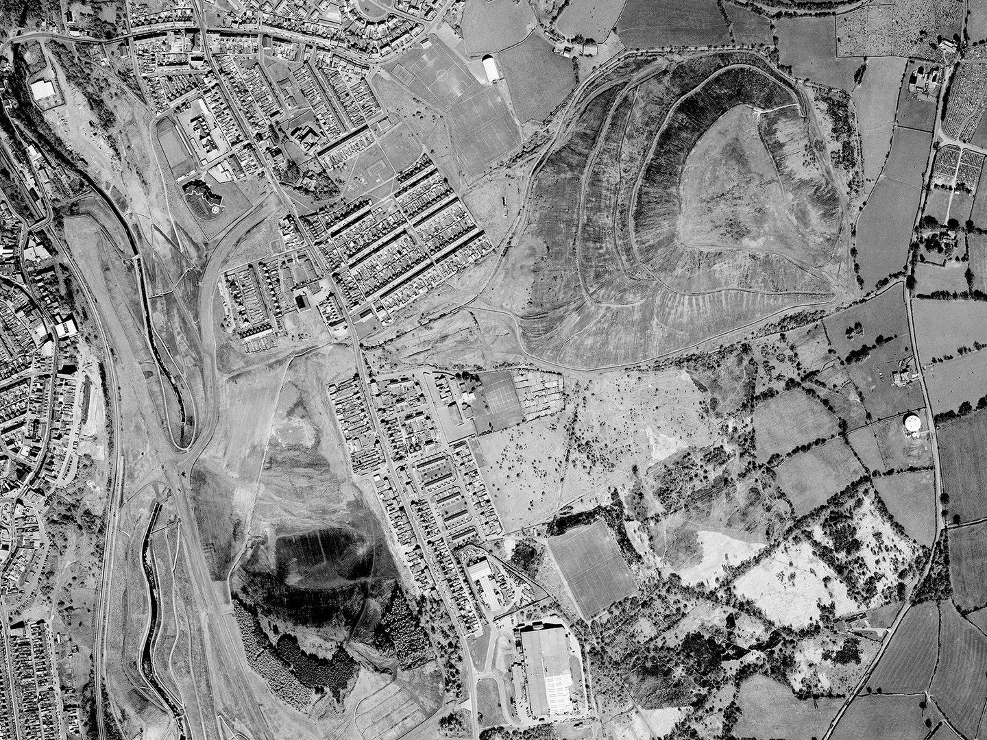 A historic black and white vertical aerial photo of a post-industrial landscape. A river runs along the left-hand edge of the image. A mix of housing and fields fills most of the image, along with two large coal spoil heaps at the top right and bottom left corners.