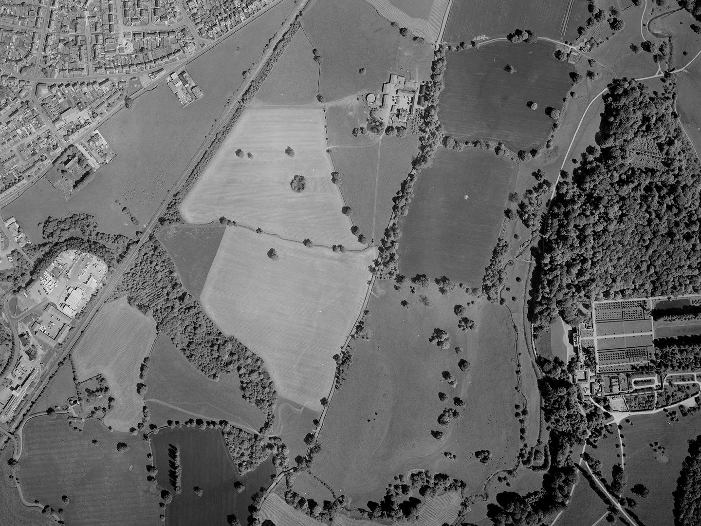 A historic black and white vertical aerial photo of fields interspersed with trees. Houses are visible in the top left corner of the image and industrial buildings at the left edge. There is a woodland to the right along with a grand stately home and its formal gardens.
