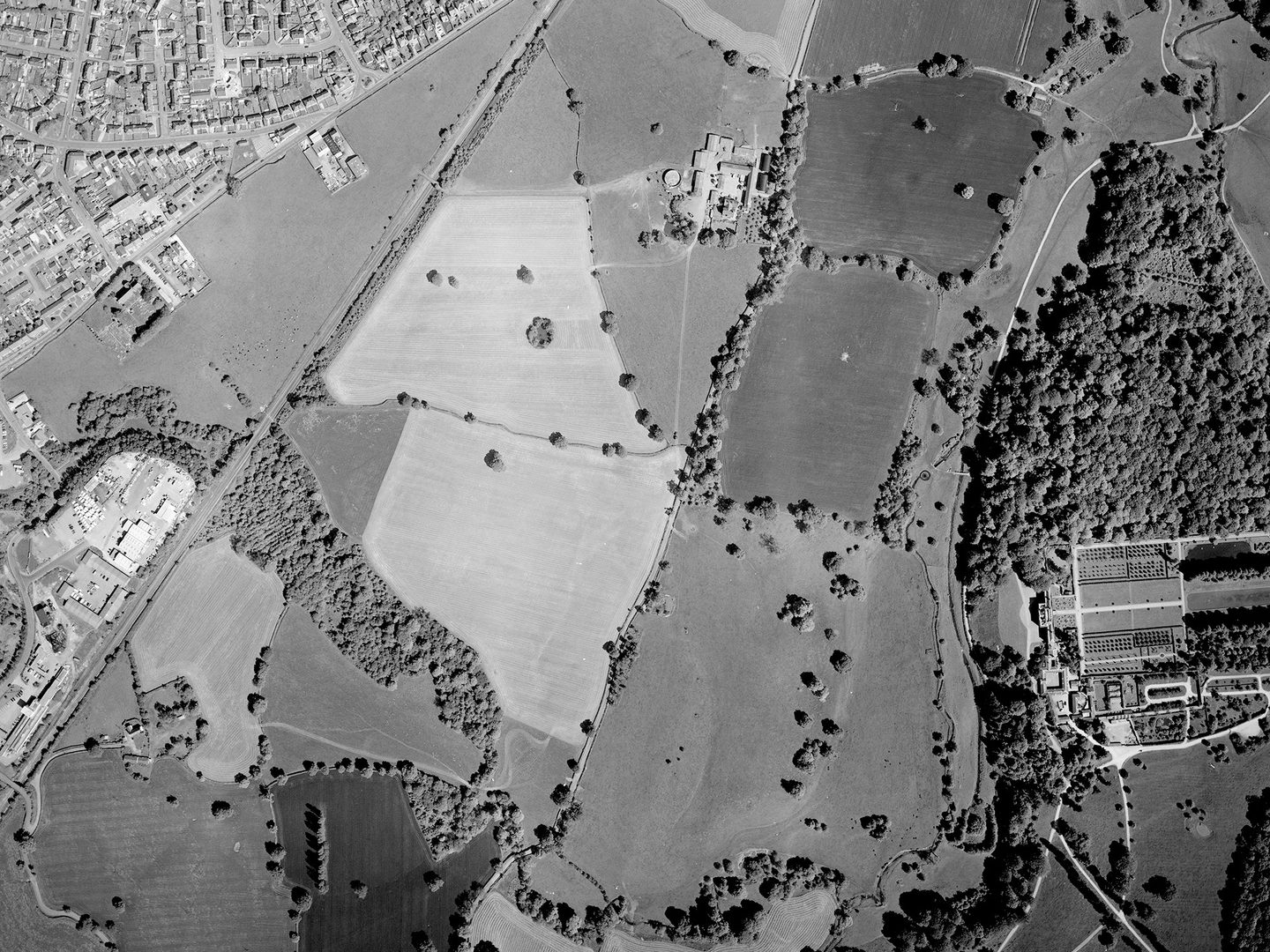 A historic black and white vertical aerial photo of fields interspersed with trees. Houses are visible in the top left corner of the image and industrial buildings at the left edge. There is a woodland to the right along with a grand stately home and its formal gardens.