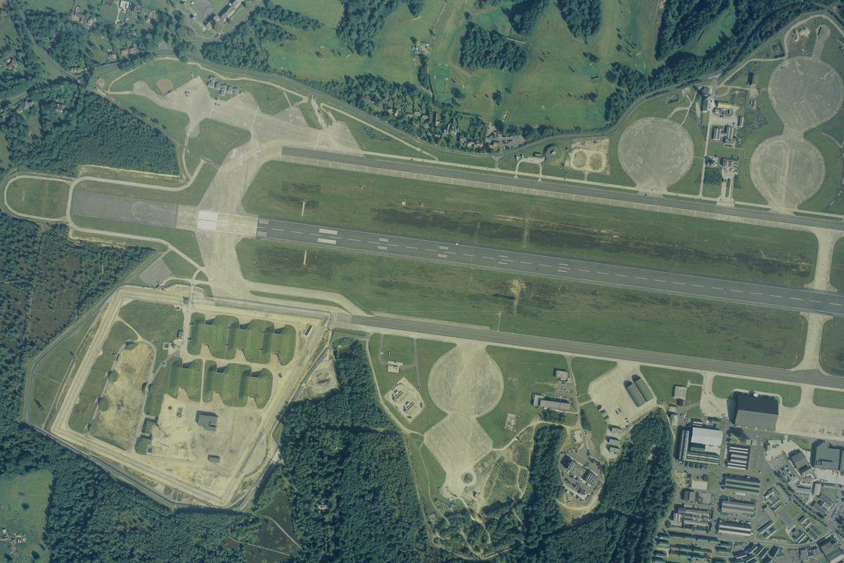 A colour aerial photo of an airfield, with a runway running horizontally across the image. The airfield is surrounded by trees. 6 grass-covered bunkers are to the lower left of the runway, with airfield buildings to the lower right.