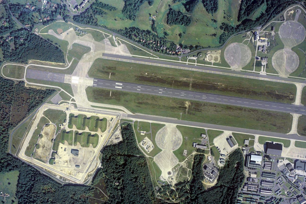 A colour aerial photo of an airfield, with a runway running horizontally across the image. The airfield is surrounded by trees. 6 grass-covered bunkers are to the lower left of the runway, with airfield buildings to the lower right.