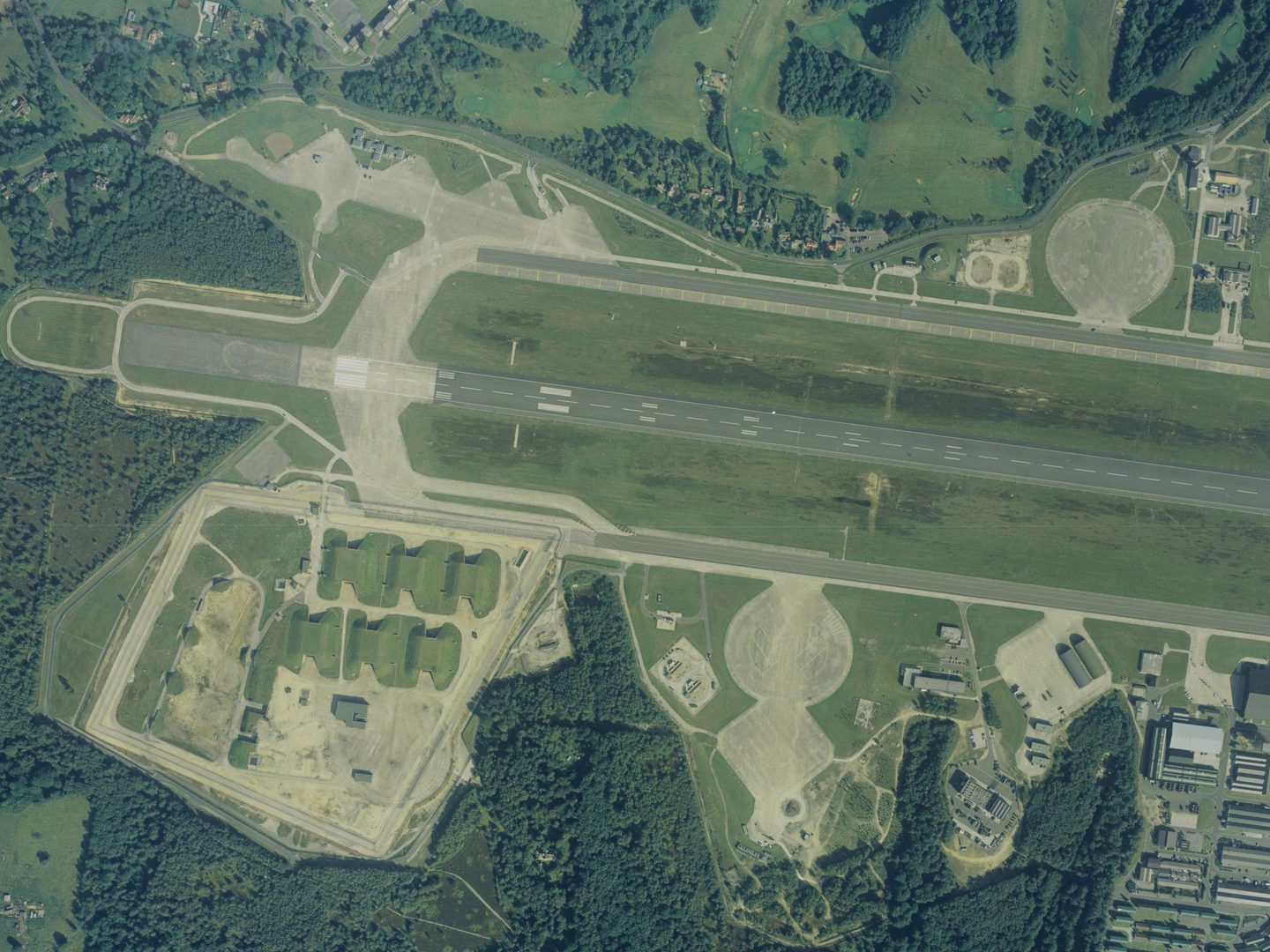 A colour aerial photo of an airfield, with a runway running horizontally across the image. The airfield is surrounded by trees. 6 grass-covered bunkers are to the lower left of the runway, with airfield buildings to the lower right.