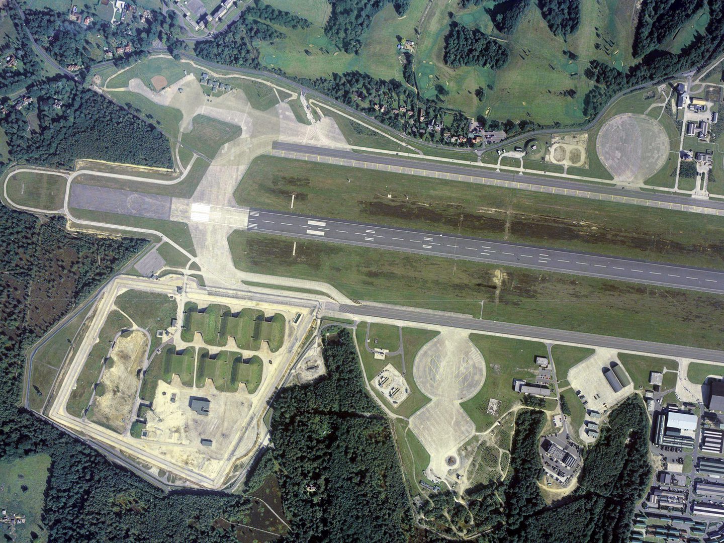 A colour aerial photo of an airfield, with a runway running horizontally across the image. The airfield is surrounded by trees. 6 grass-covered bunkers are to the lower left of the runway, with airfield buildings to the lower right.