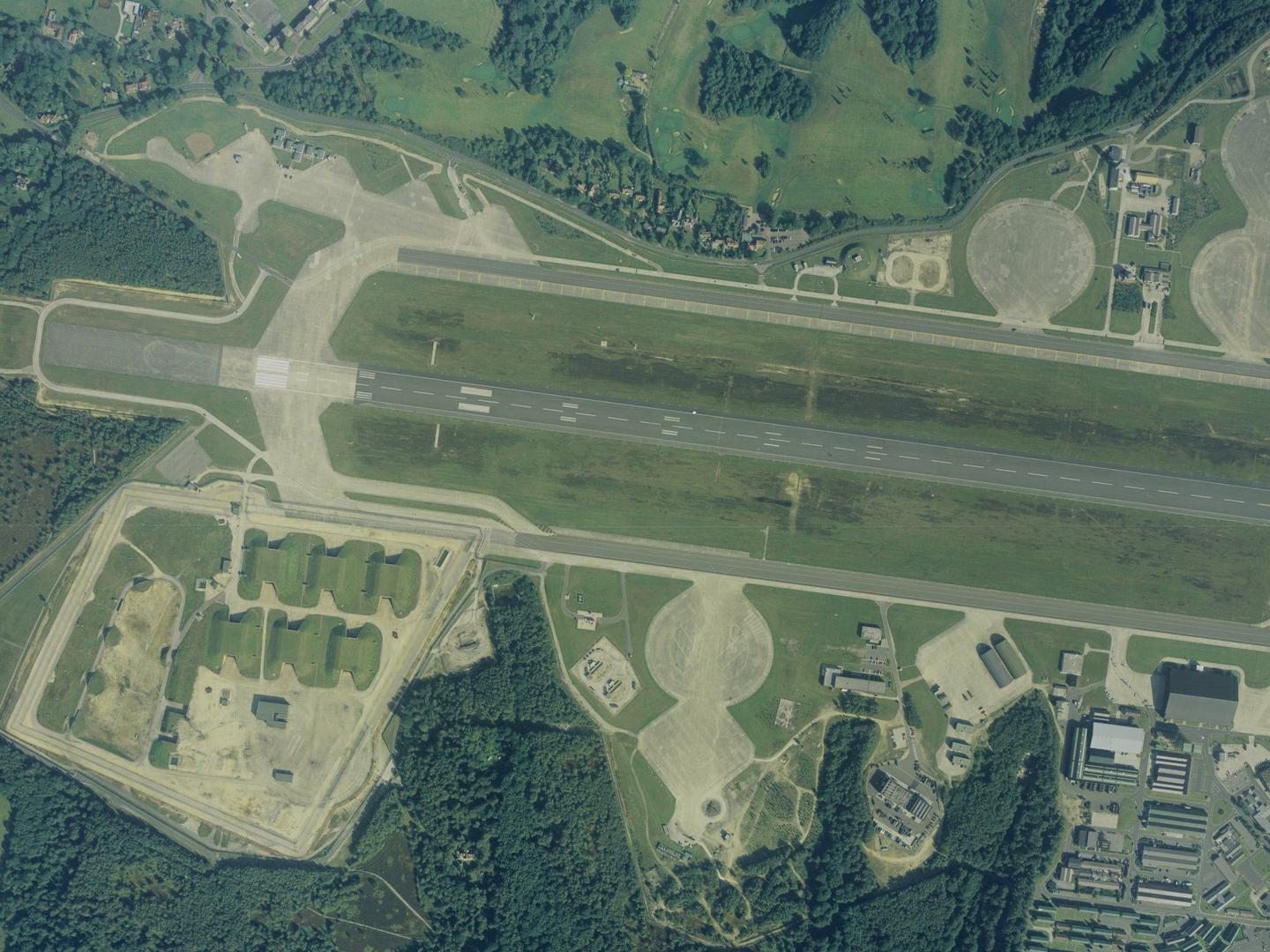 A colour aerial photo of an airfield, with a runway running horizontally across the image. The airfield is surrounded by trees. 6 grass-covered bunkers are to the lower left of the runway, with airfield buildings to the lower right.