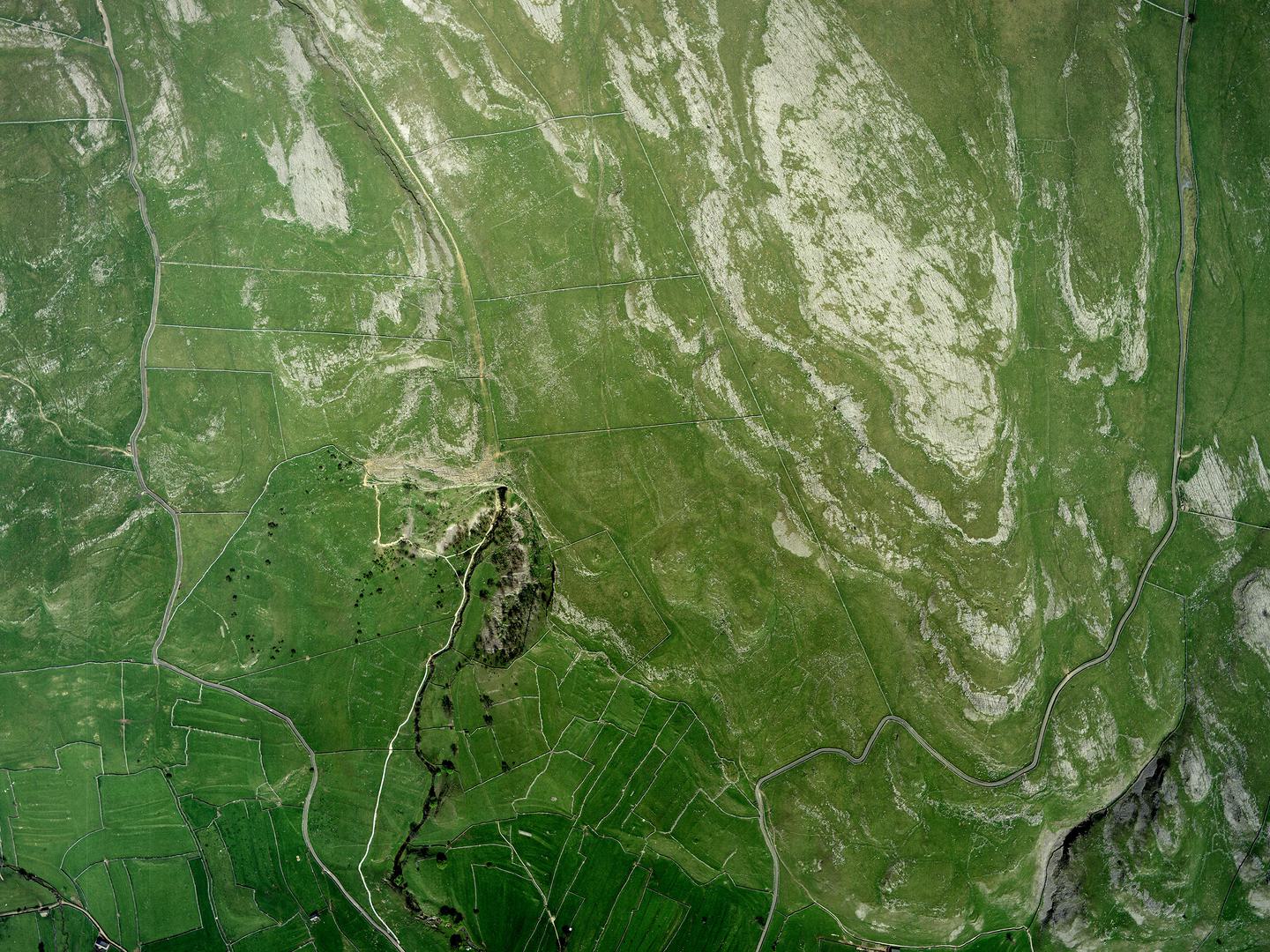 A colour aerial photo of a green landscape. The landscape is crisscrossed with a network of drystone walls. A river runs from the bottom edge to a cluster of trees in the middle of the image. Large areas of grey rock stand out against the green.