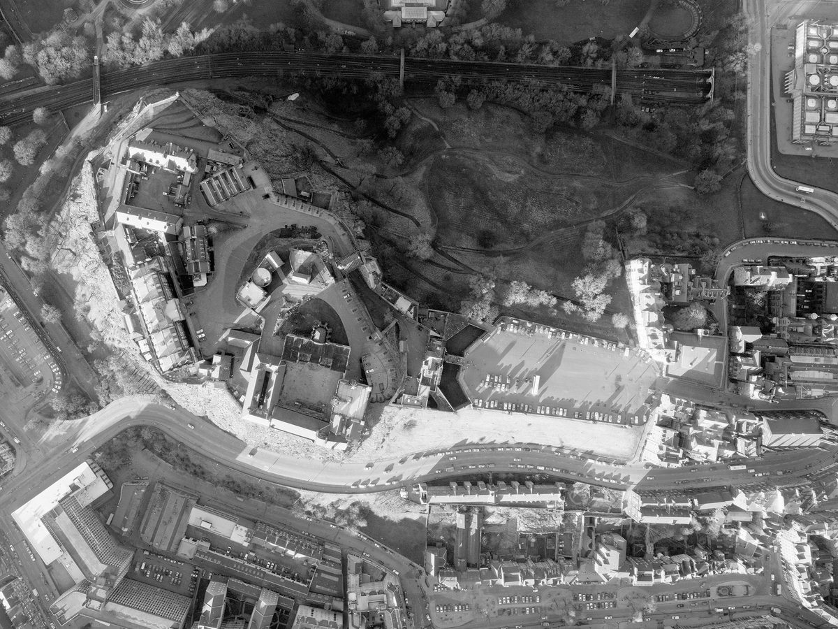 A black and white vertical aerial photo of Edinburgh Castle. The castle complex is elevated on a crag to the left of centre. A carpark and street run away from it to the right. North of the castle is a trainline and a park crossed by paths.