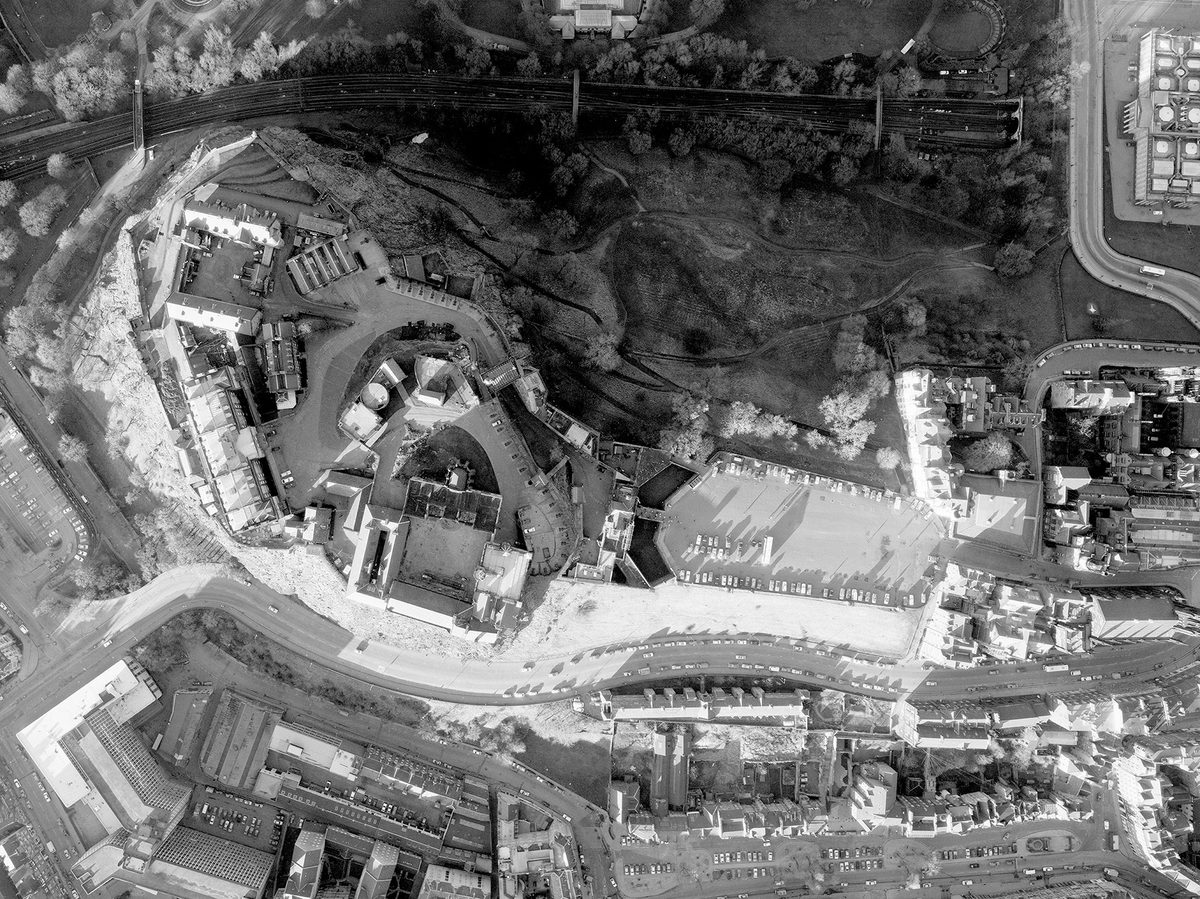 A black and white vertical aerial photo of Edinburgh Castle. The castle complex is elevated on a crag to the left of centre. A carpark and street run away from it to the right. North of the castle is a trainline and a park crossed by paths.