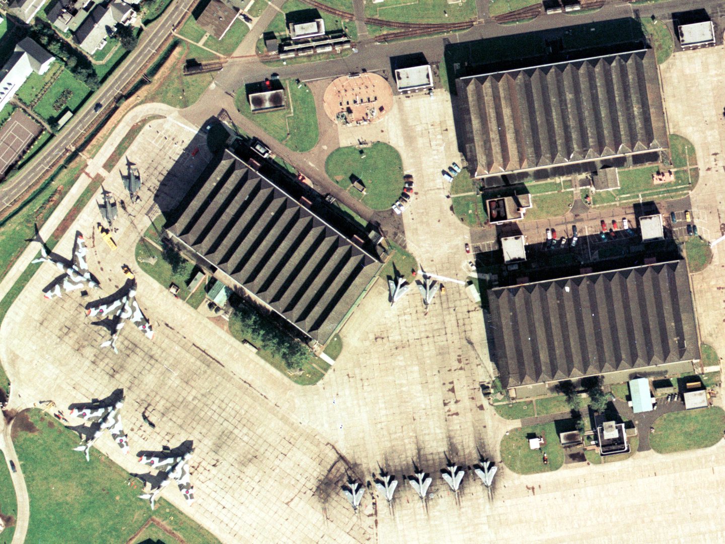 A historic colour aerial photo of an airfield with 3 large rectangular aircraft hangars. 4 Handley Page Victors painted with camouflage stripes are to the left, with 7 natural metal English Electric Lightnings in the centre. There are smaller buildings and vehicles dotted about.