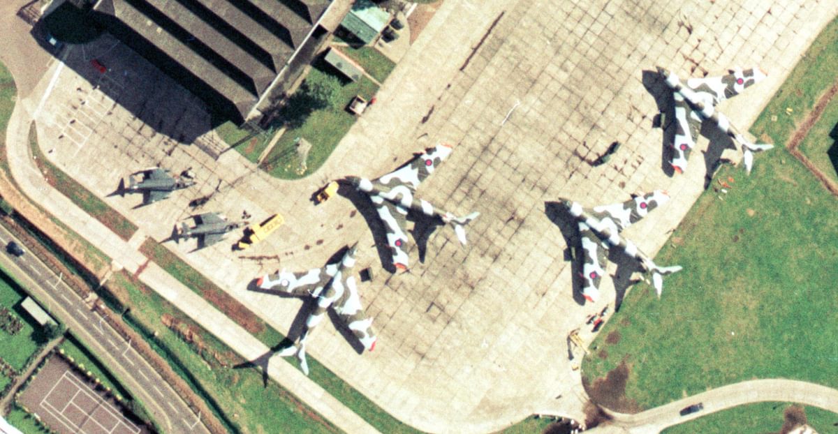 A colour aerial photo of an airfield. 4 large planes painted with camouflage stripes are to the right, with 2 smaller planes painted grey to the left. A hangar is visible at the top of the frame and vehicles are dotted about.