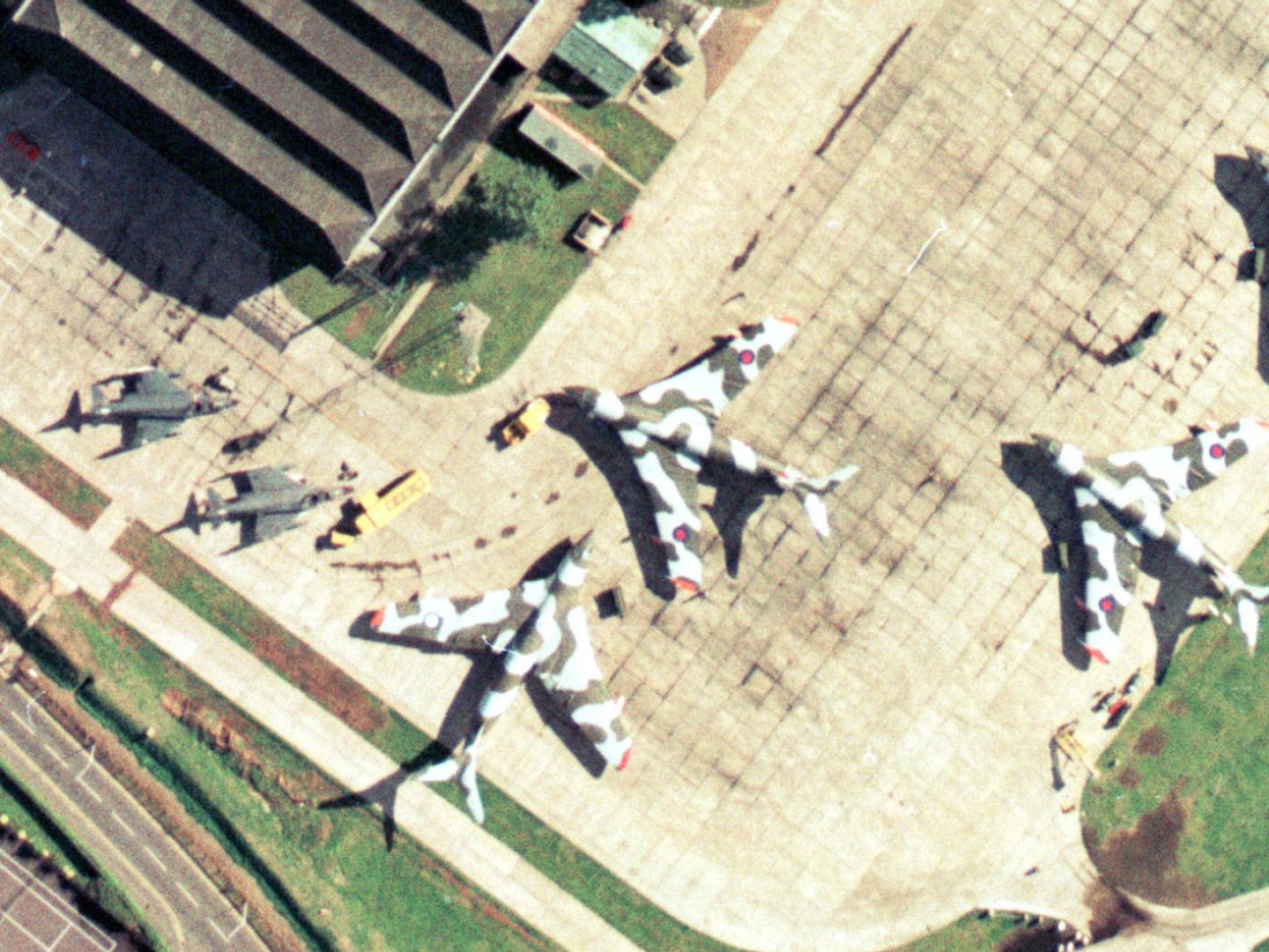 A colour aerial photo of an airfield. 4 Handley Page Victors painted with camouflage stripes are to the right, with 2 McDonnell-Douglas Phantoms painted grey to the left. A hangar is visible at the top of the frame and vehicles are dotted about.