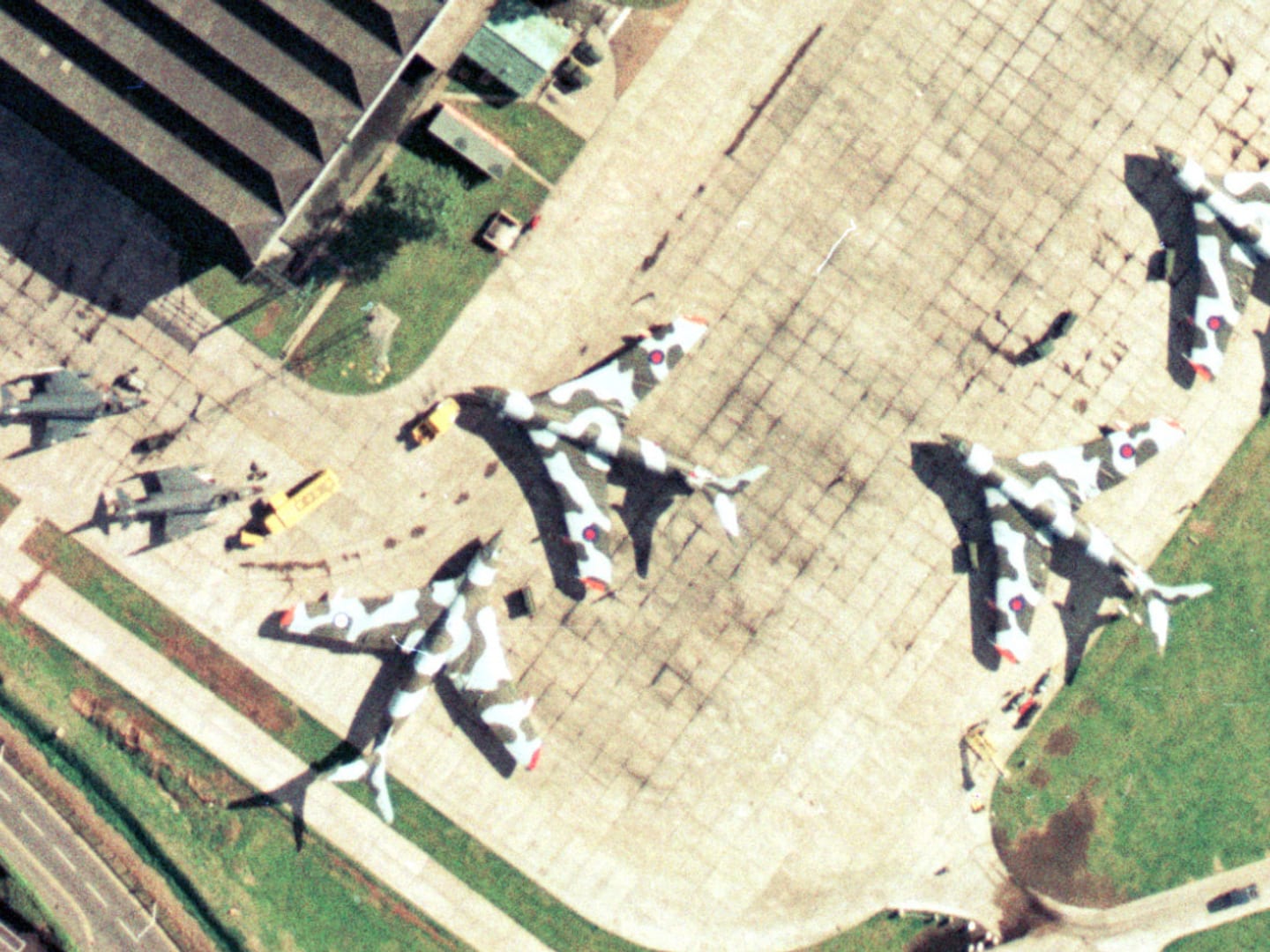 A colour aerial photo of an airfield. 4 large planes painted with camouflage stripes are to the right, with 2 smaller planes painted grey to the left. A hangar is visible at the top of the frame and vehicles are dotted about.