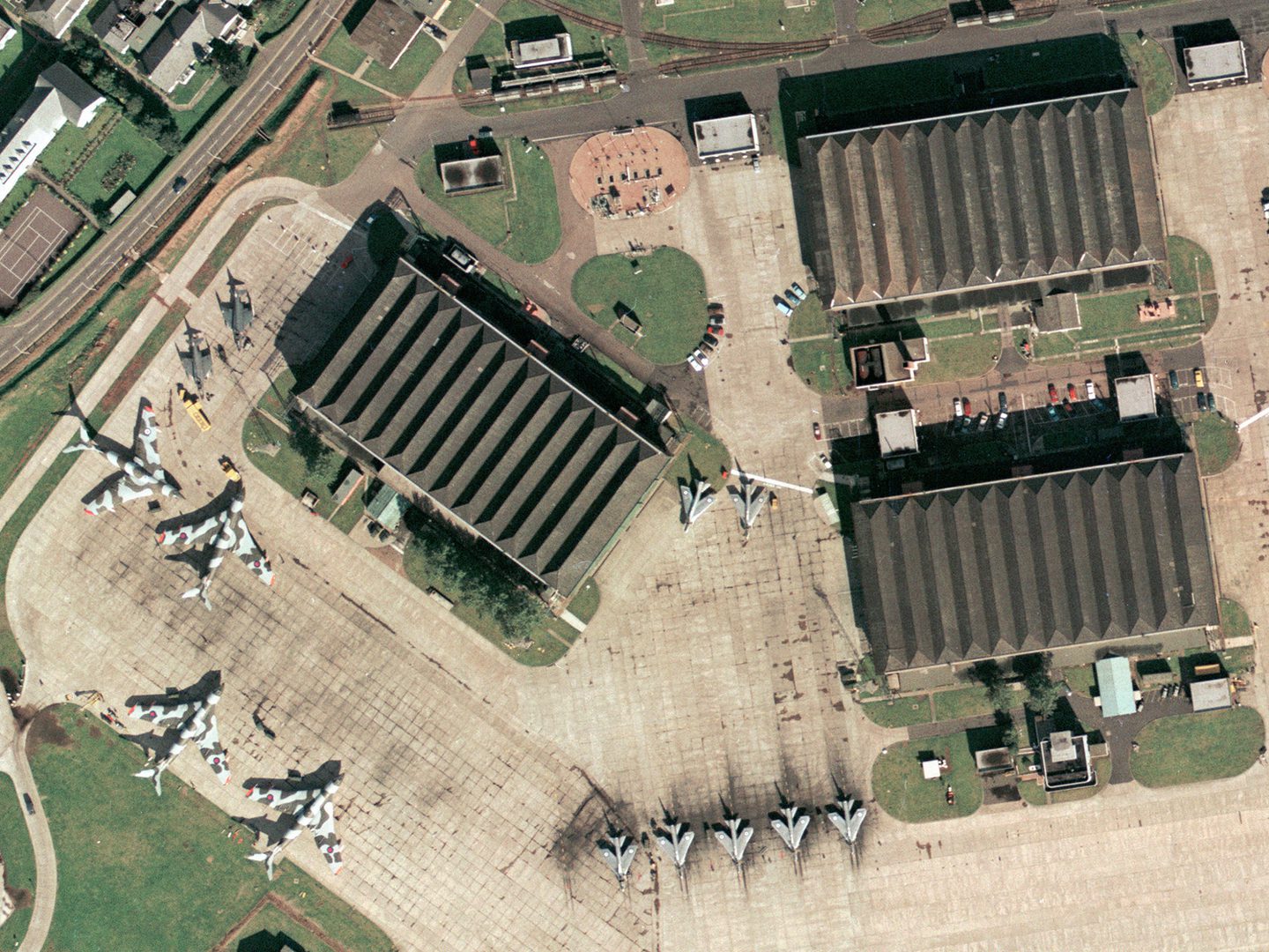 A historic colour aerial photo of an airfield with 3 large rectangular aircraft hangars. 4 Handley Page Victors painted with camouflage stripes are to the left, with 7 natural metal English Electric Lightnings in the centre. There are smaller buildings and vehicles dotted about.