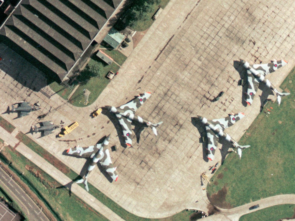 A colour aerial photo of an airfield. 4 Handley Page Victors painted with camouflage stripes are to the right, with 2 McDonnell-Douglas Phantoms painted grey to the left. A hangar is visible at the top of the frame and vehicles are dotted about.
