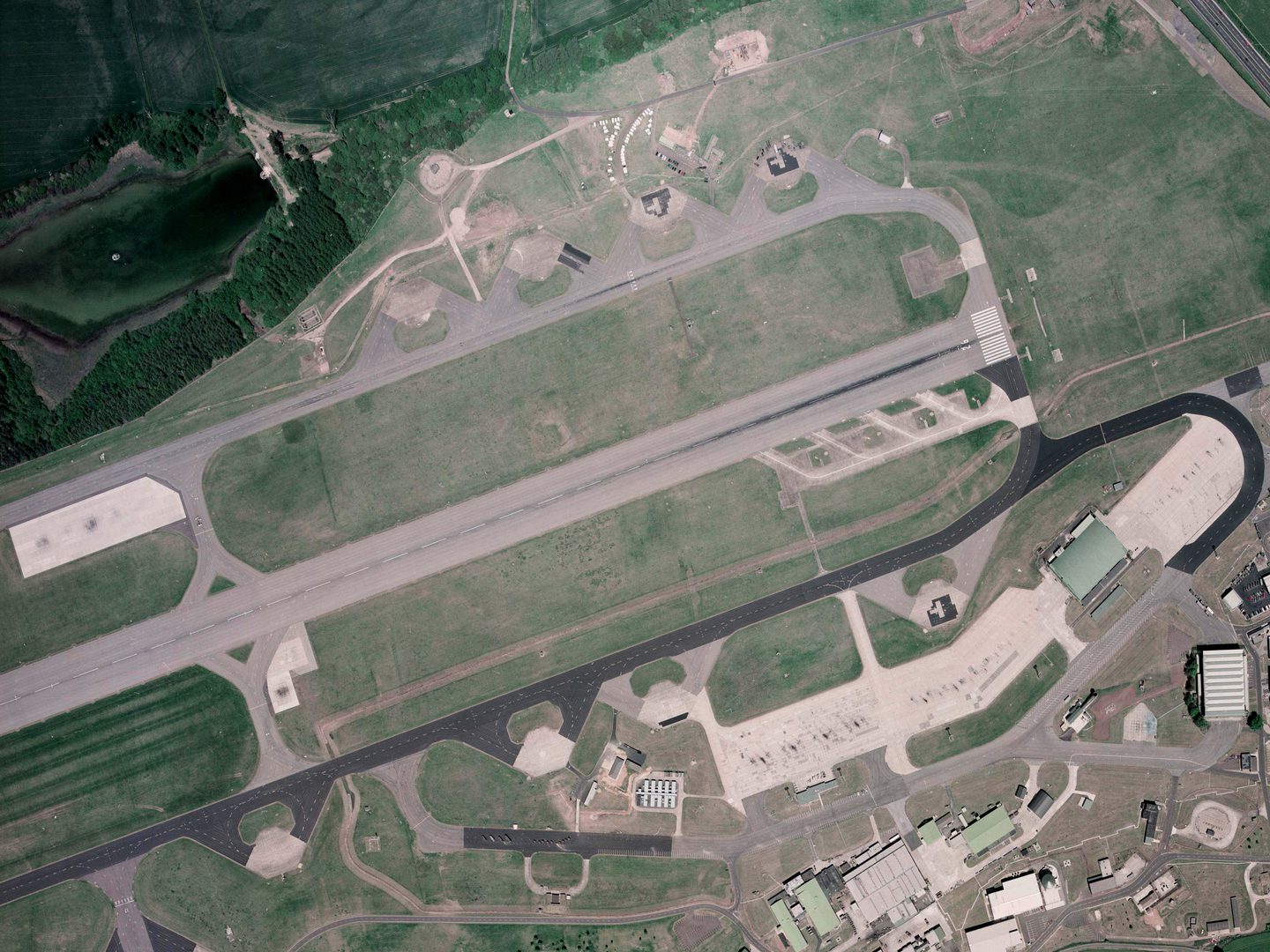 A colour aerial photo of an airfield. A runway flanked by taxiways runs horizontally across the middle of the image. At the right-hand end of the runway are 4 small concrete platforms. There are airport buildings bottom right and trees at the top.