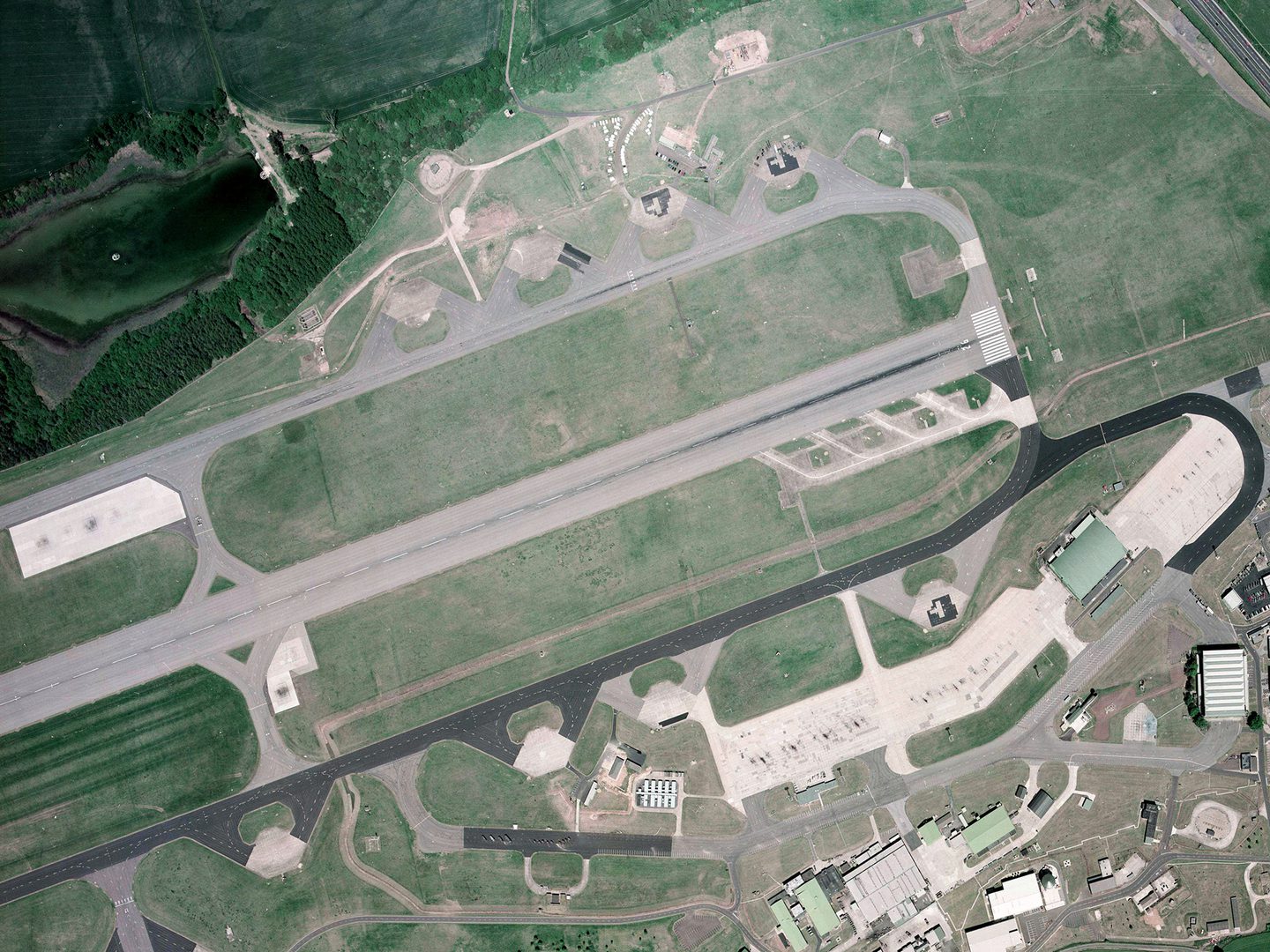 A colour aerial photo of an airfield. A runway flanked by taxiways runs horizontally across the middle of the image. At the right-hand end of the runway are 4 small concrete platforms. There are airport buildings bottom right and trees at the top.