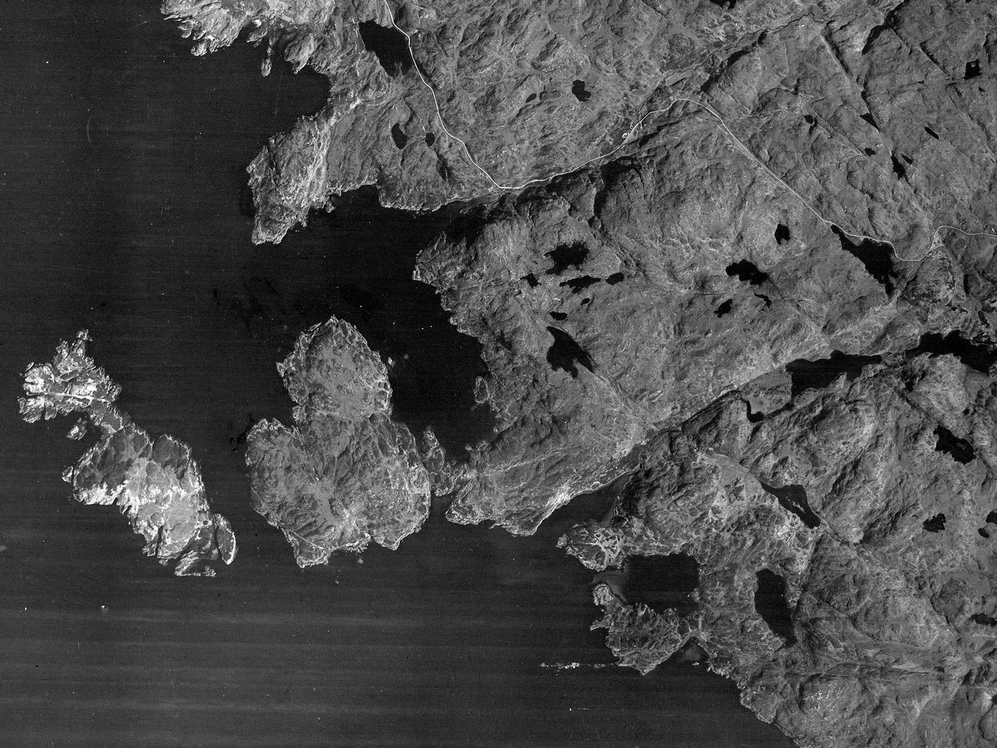 A historic black and white vertical vertical aerial photo of a coastline. The land to the right is dotted with numerous bodies of water and has a ragged edge where it meets the sea. Two islands project into the sea to the left.