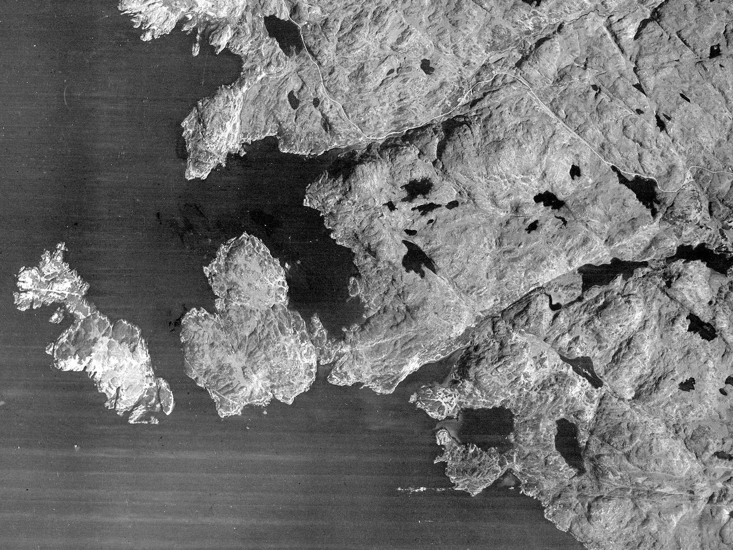 A historic black and white vertical vertical aerial photo of a coastline. The land to the right is dotted with numerous bodies of water and has a ragged edge where it meets the sea. Two islands project into the sea to the left.