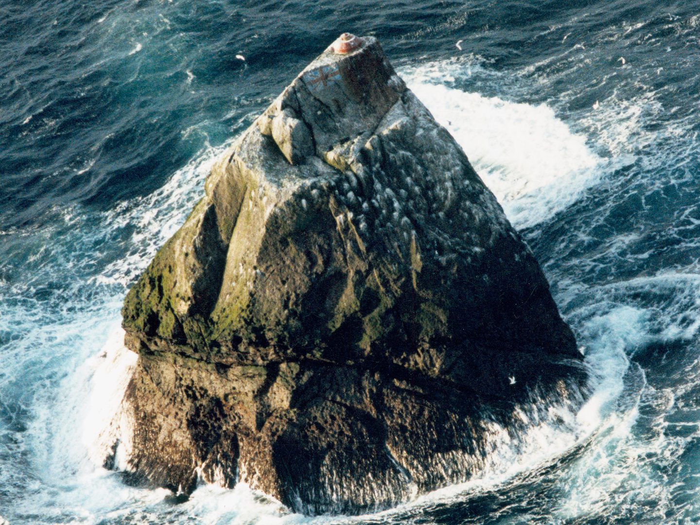 A historic colour oblique aerial photo of Rockall - a triangular shaped rock surrounded by blue sea and white foam. On the peak of the rock is a small orange dome next to a painted Union Flag.