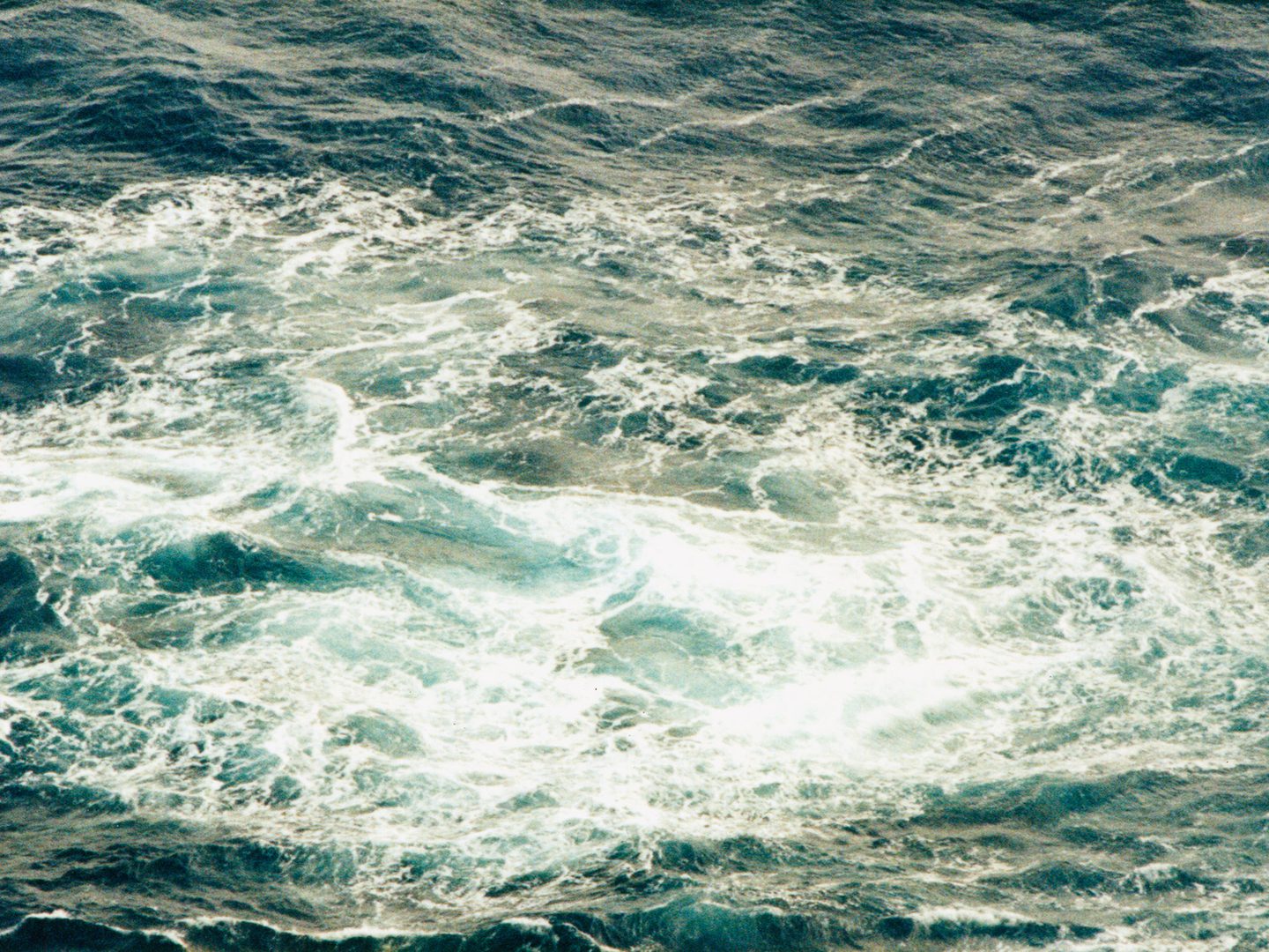 A historic colour oblique aerial photo of white foam on the surface of the sea.