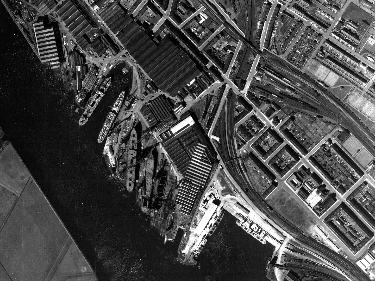 A historic black and white vertical aerial photo of an area of dockland. A river runs from the top left corner of the image to the bottom centre. The docks are to the right, with several ships moored at them. Various industrial buildings cover the shore.