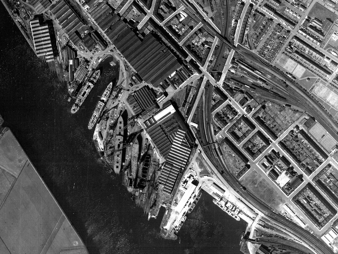 A historic black and white vertical aerial photo of an area of dockland. A river runs from the top left corner of the image to the bottom centre. The docks are to the right, with several ships moored at them. Various industrial buildings cover the shore.
