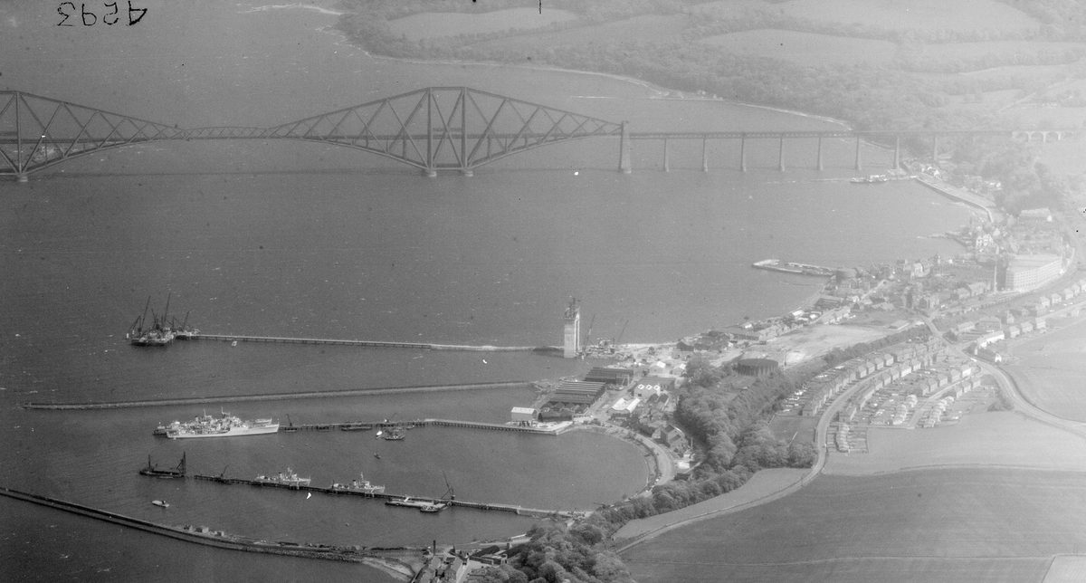 A historic black and white oblique aerial photo of the Forth Rail Bridge stretching across the Firth of Forth. The bridge is formed of iron girders arranged in rhomboid shapes. In the foreground is a small town with jetties reaching into the Forth.