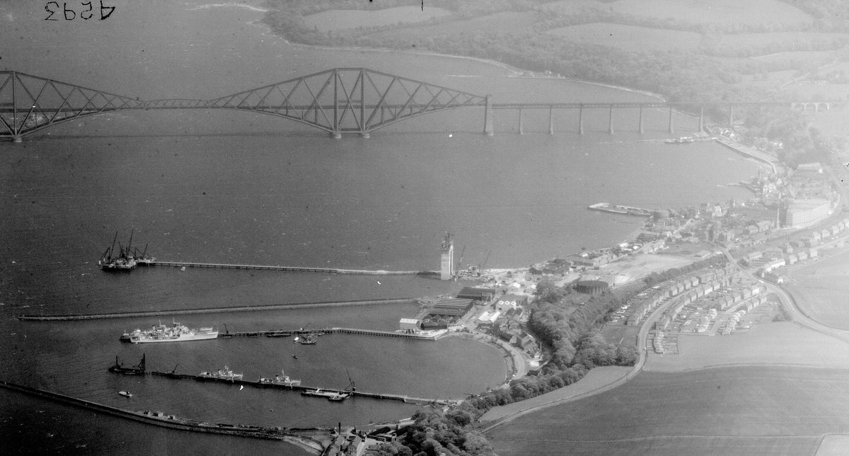 A historic black and white oblique aerial photo of the Forth Rail Bridge stretching across the Firth of Forth. The bridge is formed of iron girders arranged in rhomboid shapes. In the foreground is a small town with jetties reaching into the Forth.