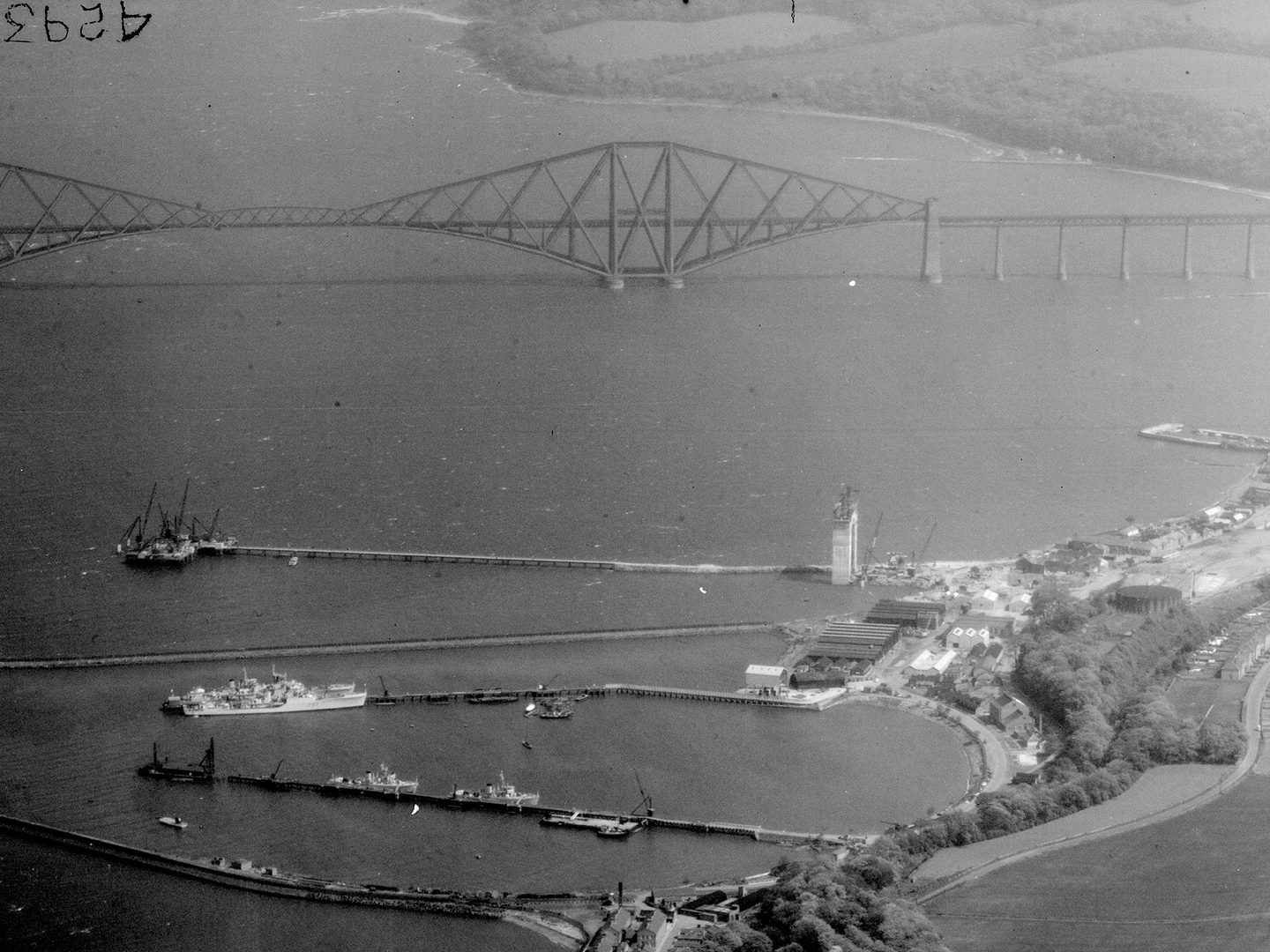 A historic black and white oblique aerial photo of the Forth Rail Bridge stretching across the Firth of Forth. The bridge is formed of iron girders arranged in rhomboid shapes. In the foreground is a small town with jetties reaching into the Forth.