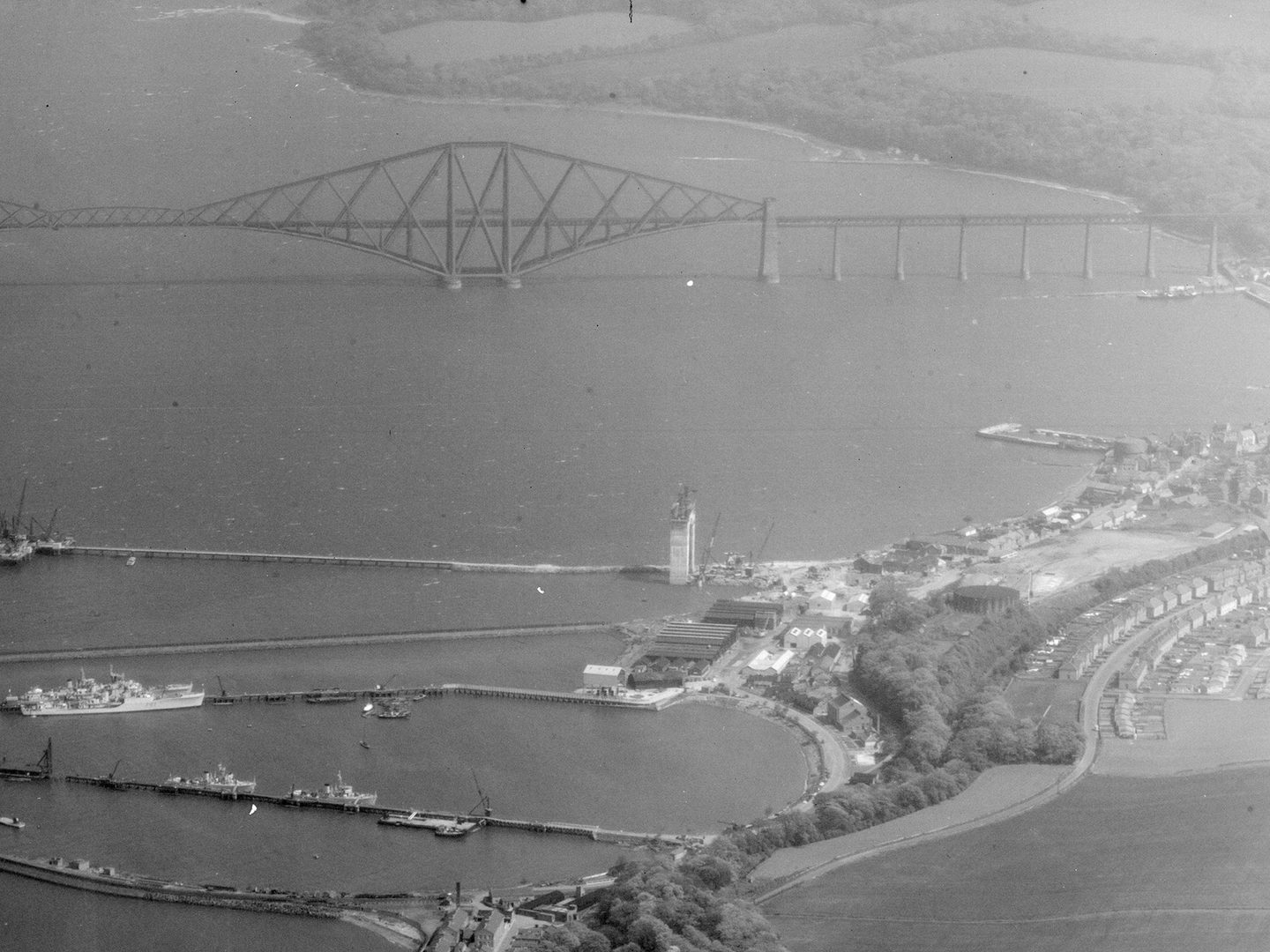 A historic black and white oblique aerial photo of the Forth Rail Bridge stretching across the Firth of Forth. The bridge is formed of iron girders arranged in rhomboid shapes. In the foreground is a small town with jetties reaching into the Forth.