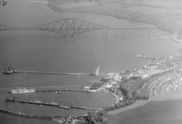 A historic black and white oblique aerial photo of the Forth Rail Bridge stretching across the Firth of Forth. The bridge is formed of iron girders arranged in rhomboid shapes. In the foreground is a small town with jetties reaching into the Forth.