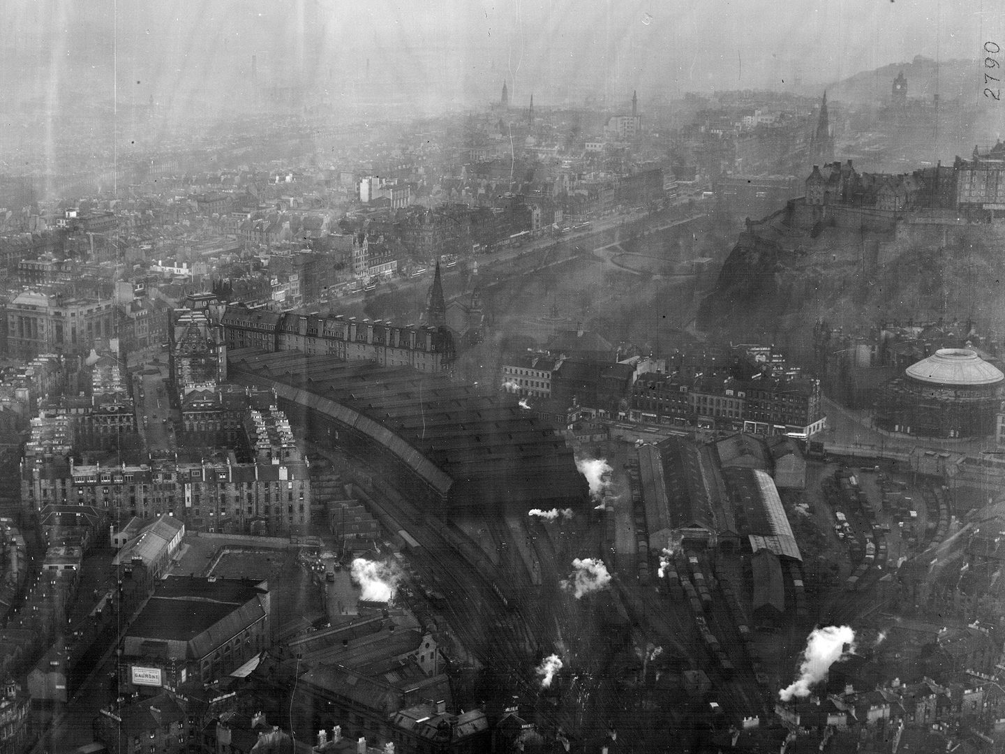 A historic black and white oblique aerial photo of Edinburgh from the west, with the Castle visible on its hill to the right of the image. The photo is quite faded and grainy.