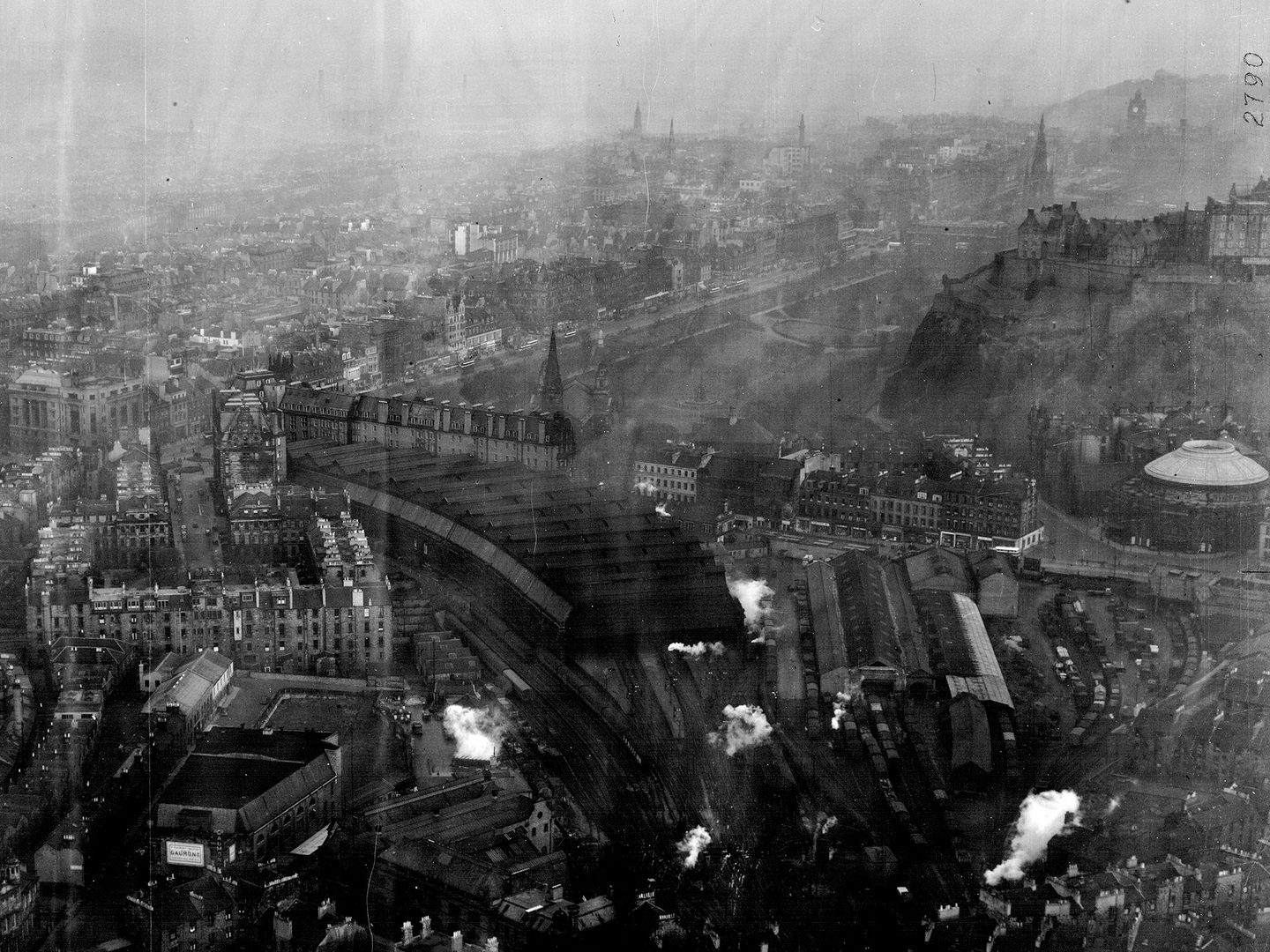 A historic black and white oblique aerial photo of Edinburgh from the west, with the Castle visible on its hill to the right of the image. The photo is quite faded and grainy.