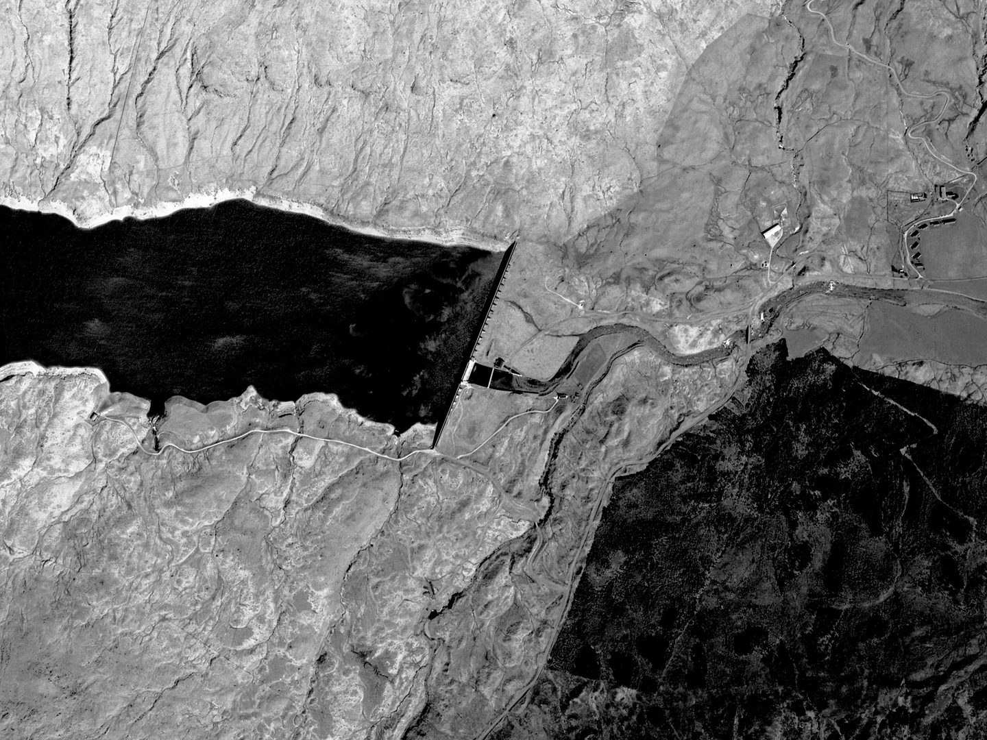 A historic black and white vertical aerial photo of a highland landscape. A loch is visible to the left, terminating in a dam running vertically through the image. A woodland is visible in the bottom right corner.