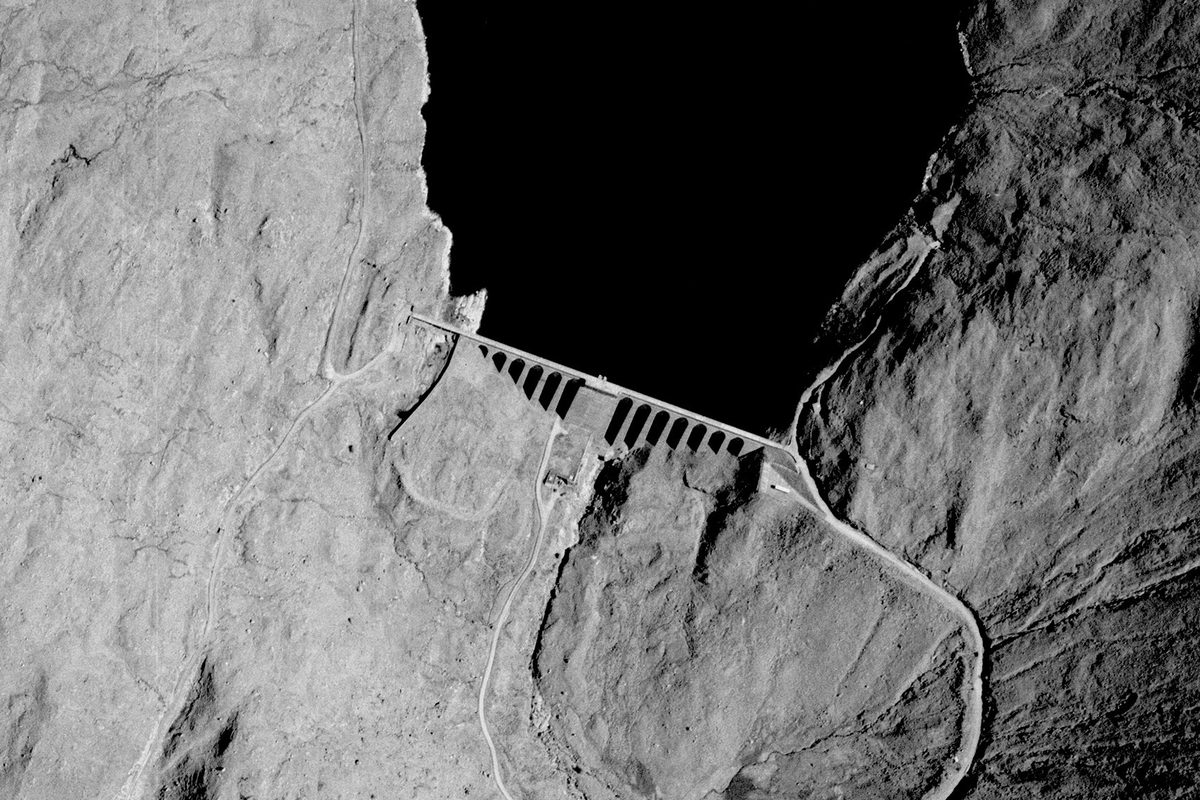 A historic black and white vertical aerial photo of a dam across the foot of a loch. The downstream face of the dam is comprised of numerous narrow arches. The loch sits in a highland landscape.