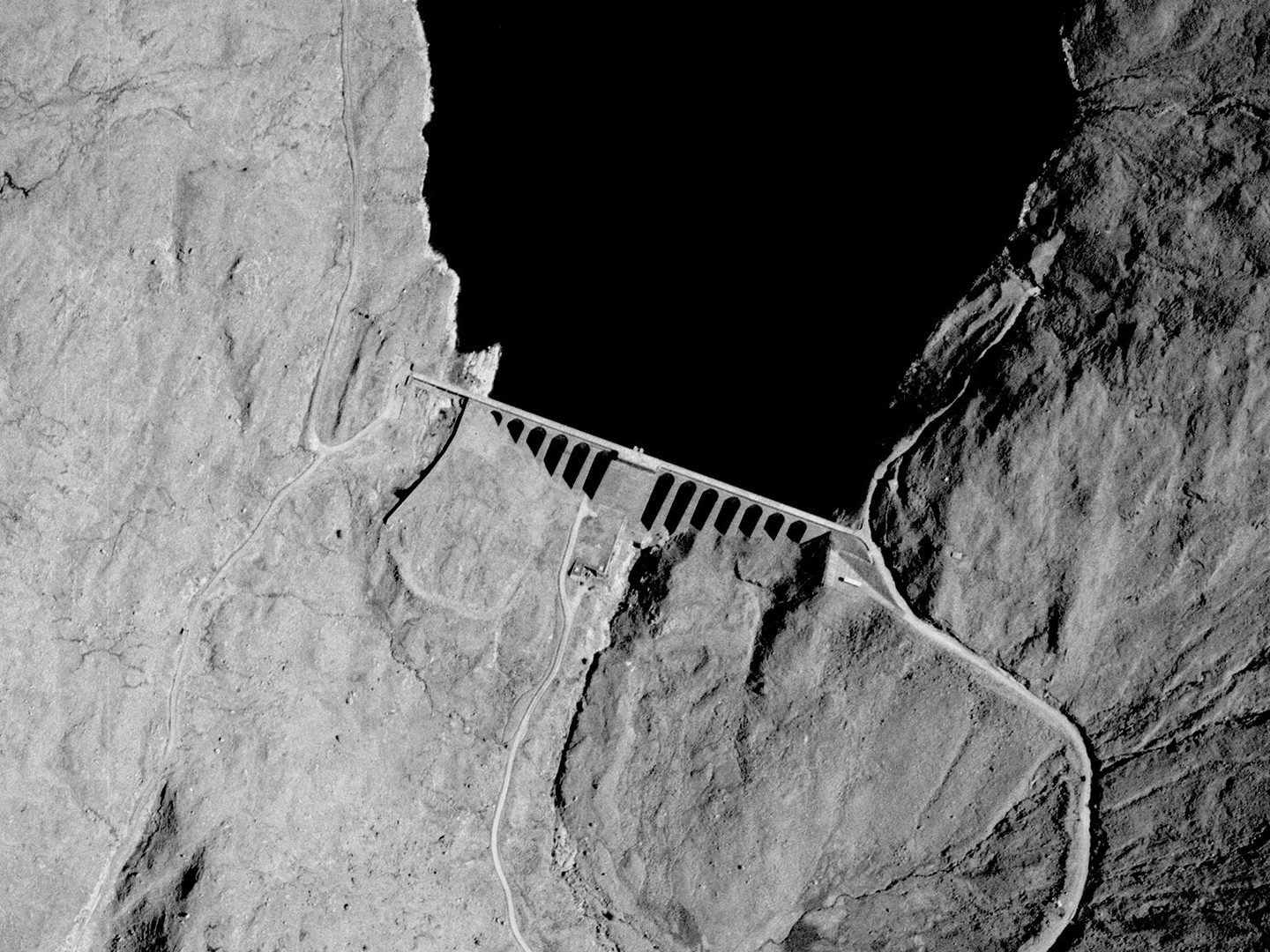 A historic black and white vertical aerial photo of a dam across the foot of a loch. The downstream face of the dam is comprised of numerous narrow arches. The loch sits in a highland landscape.