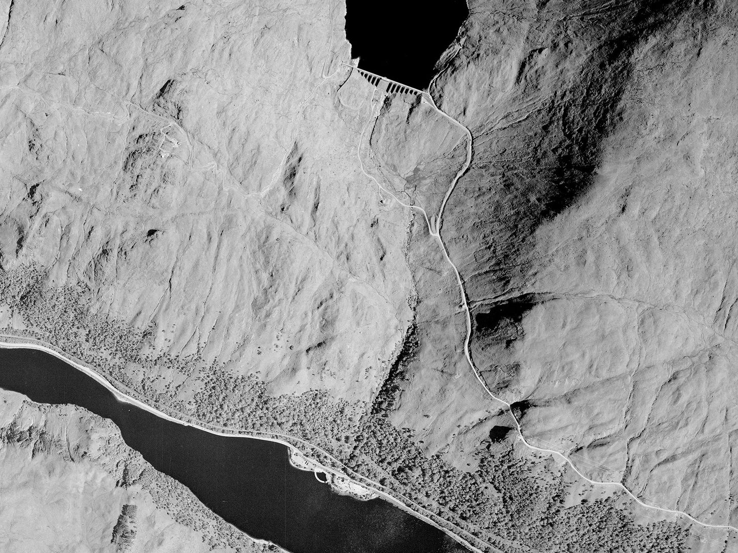 A historic black and white vertical aerial photo of a highland landscape. At the top centre edge an section of loch can be seen held back by an arched dam. Another loch runs along the bottom edge of the image, fringed with trees.