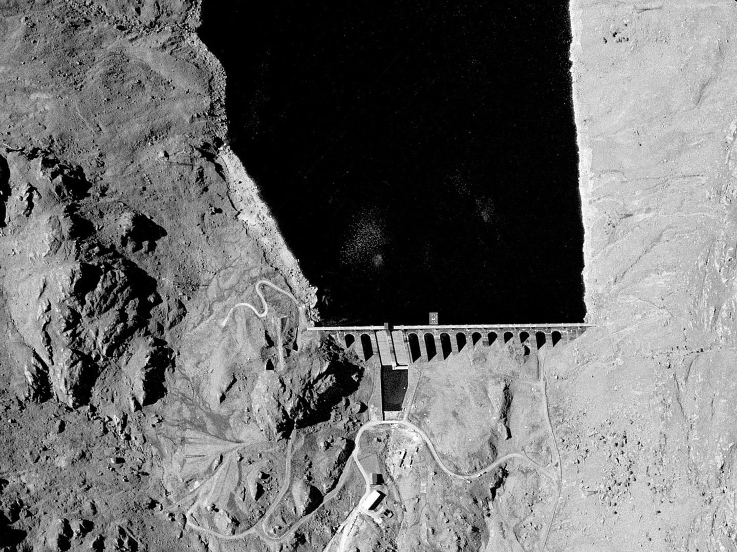 A historic black and white vertical aerial photo of a loch terminating in a dam. The downstream face of the dam is comprised of several narrow arches. There is a rocky landscape to the left of the loch.
