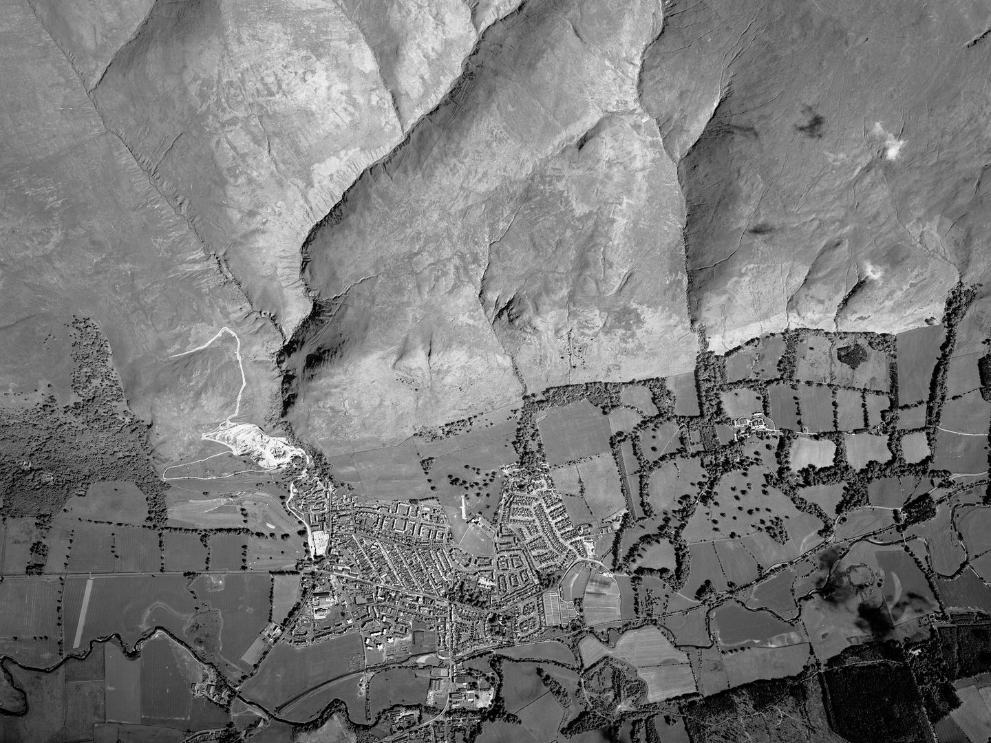 A black and white vertical aerial photo of a landscape. The top half of the image shows bare moorland, with several valleys marking the location of streams. The bottom half shows a small town surrounded by fields and scattered woodland.