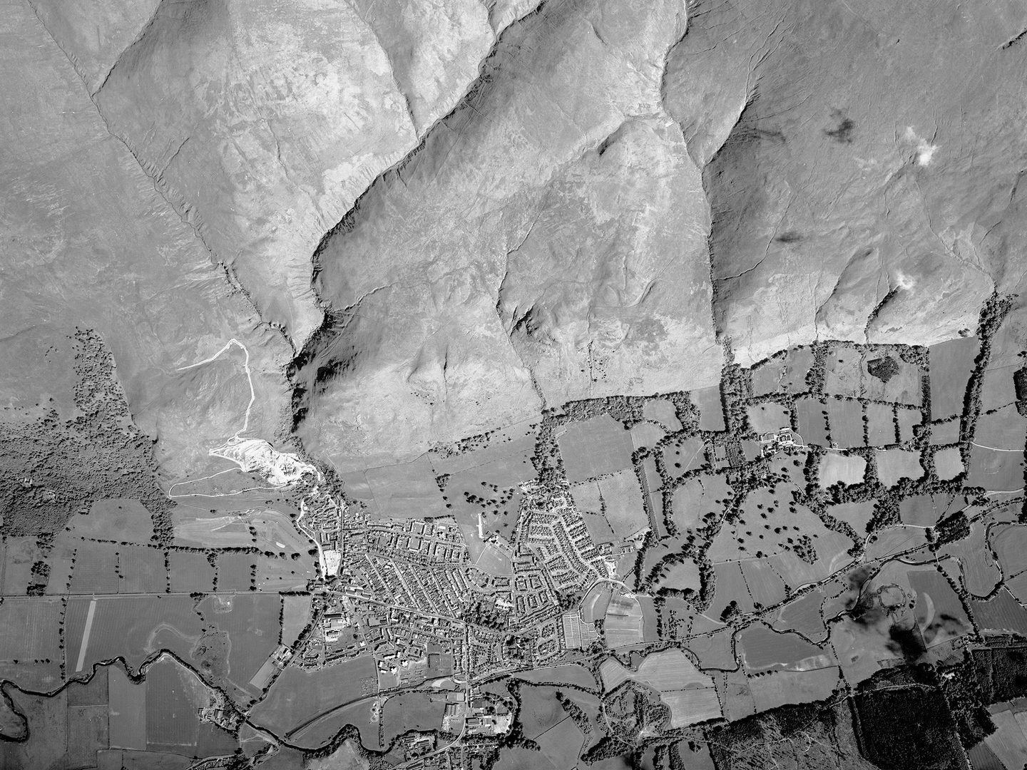 A black and white vertical aerial photo of a landscape. The top half of the image shows bare moorland, with several valleys marking the location of streams. The bottom half shows a small town surrounded by fields and scattered woodland.
