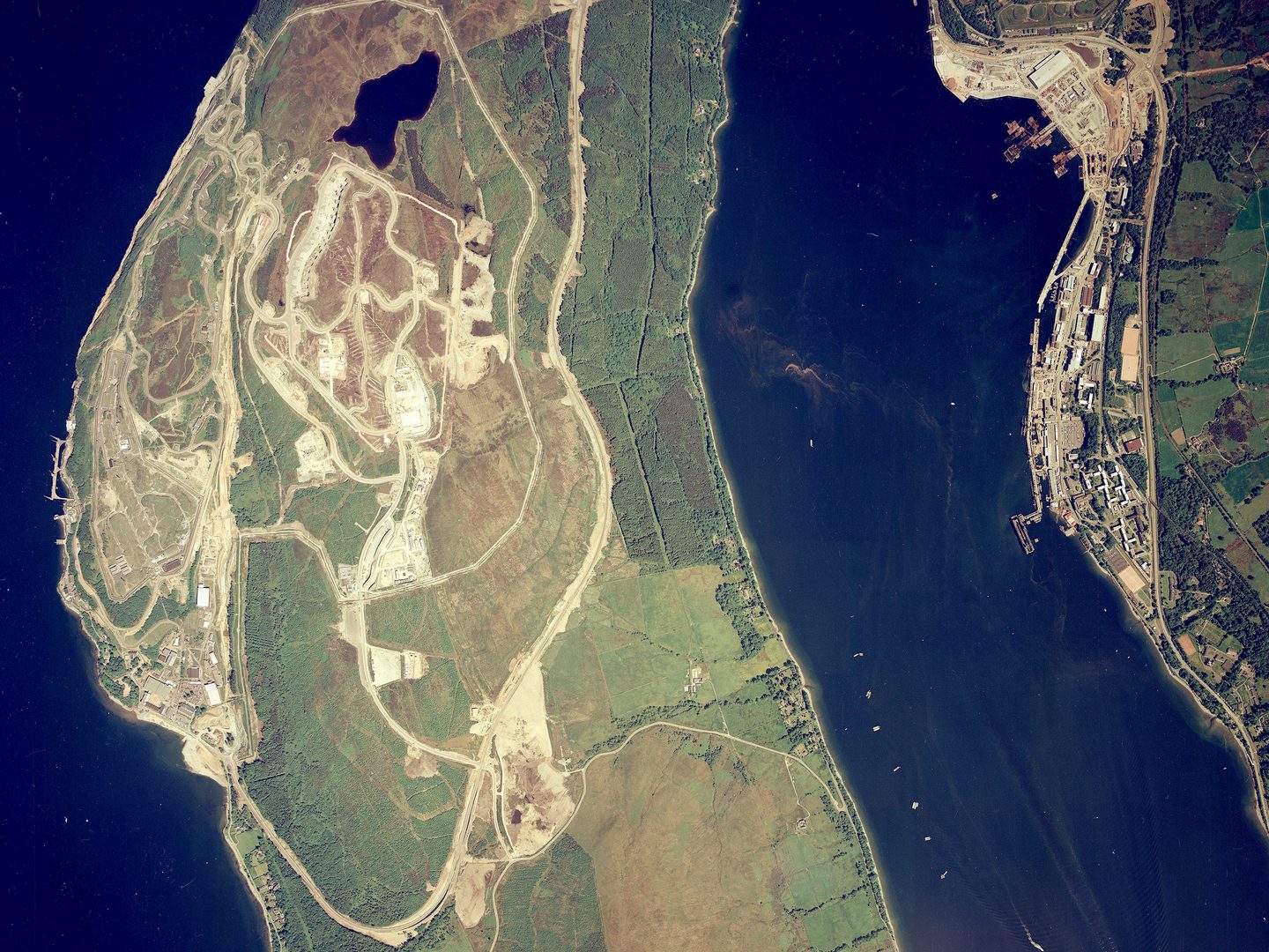 A colour aerial photo of a strip of forested land bordered on the left by a dark blue sea. There is a complex of buildings on the left-hand coast. To the right is another area of sea bordered by a 2nd area of land, on which is a large port complex.