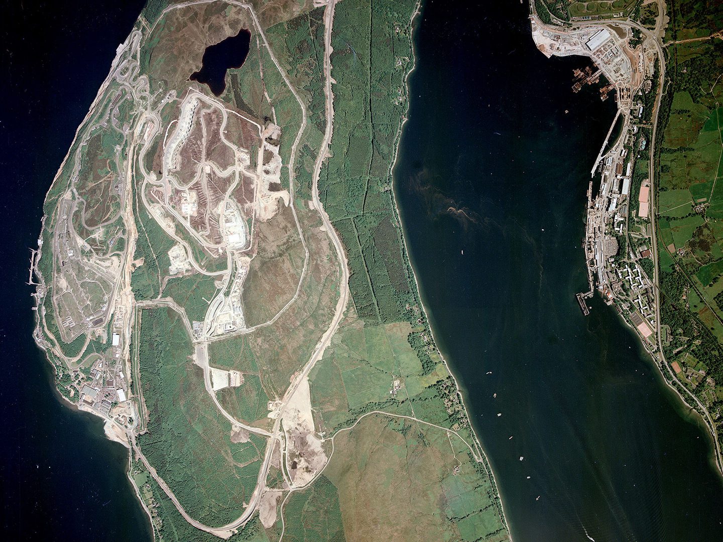 A colour aerial photo of a strip of forested land bordered on the left by a dark blue sea. There is a complex of buildings on the left-hand coast. To the right is another area of sea bordered by a 2nd area of land, on which is a large port complex.