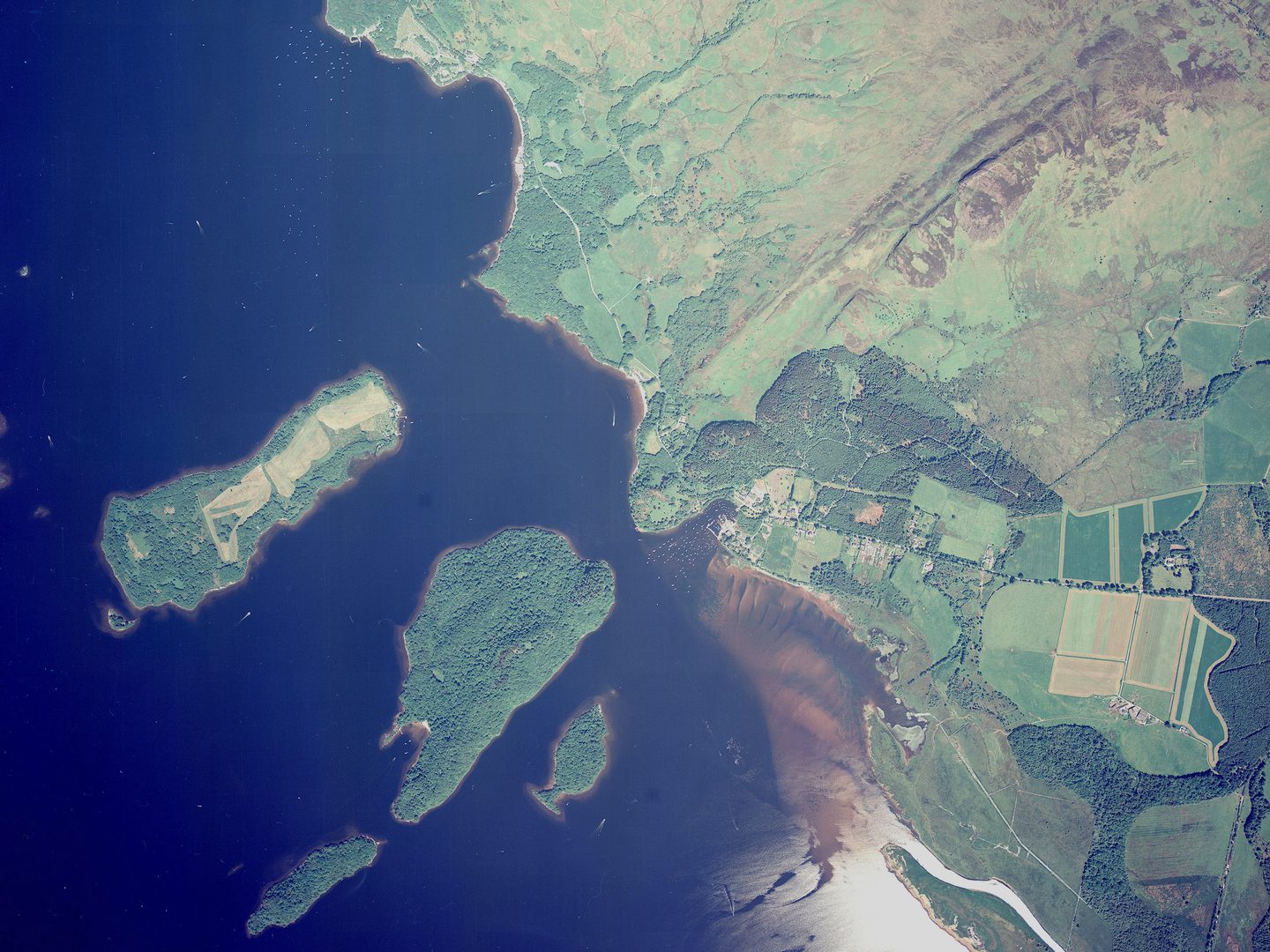 A colour vertical aerial photo of Loch Lomond. The left-hand side of the image shows the blue water of the loch, with 5 heavily wooded islands. To the right is the shore of the loch, which is mostly green moorland. There are a few woods and fields.
