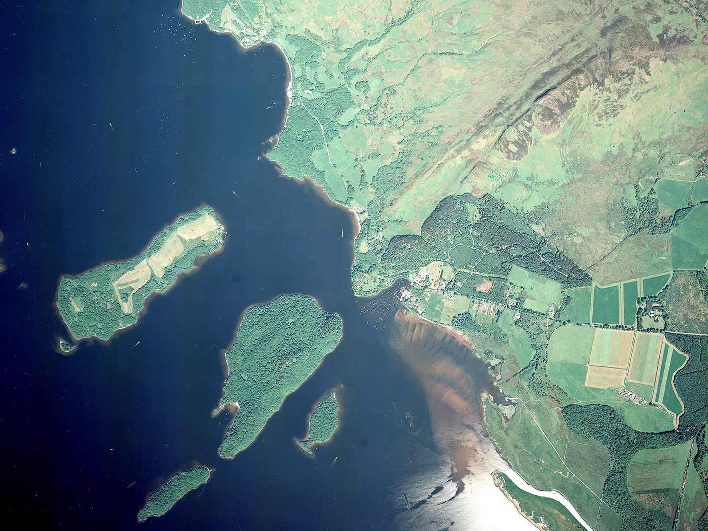 A colour vertical aerial photo of Loch Lomond. The left-hand side of the image shows the blue water of the loch, with 5 heavily wooded islands. To the right is the shore of the loch, which is mostly green moorland. There are a few woods and fields.