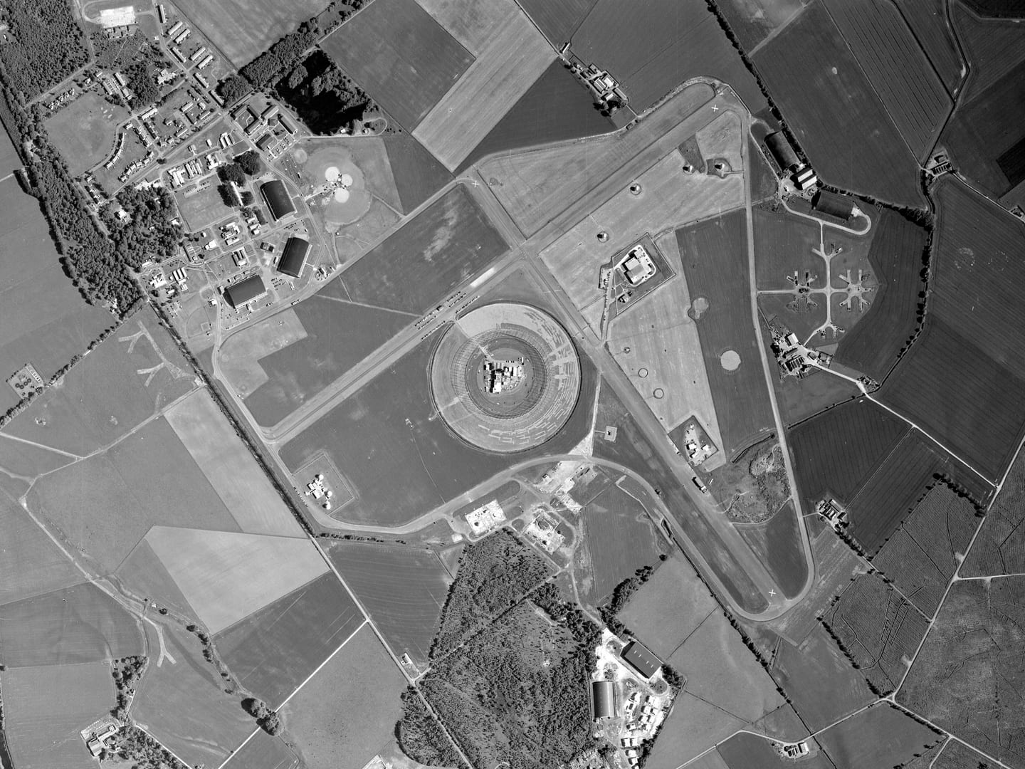 A black and white aerial photo centred on a large, circular cage-like structure. It sits in the angle of 2 runways crossing at right angles. The runways are surrounded by smaller structures. There is a village top left and a small wood bottom centre.