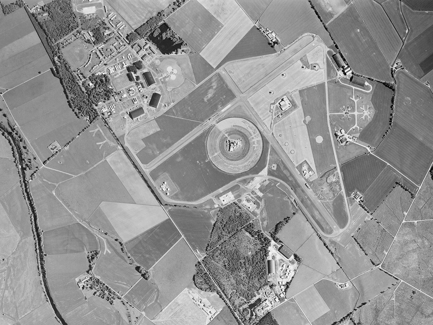 A historic black and white vertical aerial photo of an airfield. 2 runways cross in an X shape in the middle of the image. In the angle of the cross is a circular enclosure, and 3 hangars are visible to the top left.