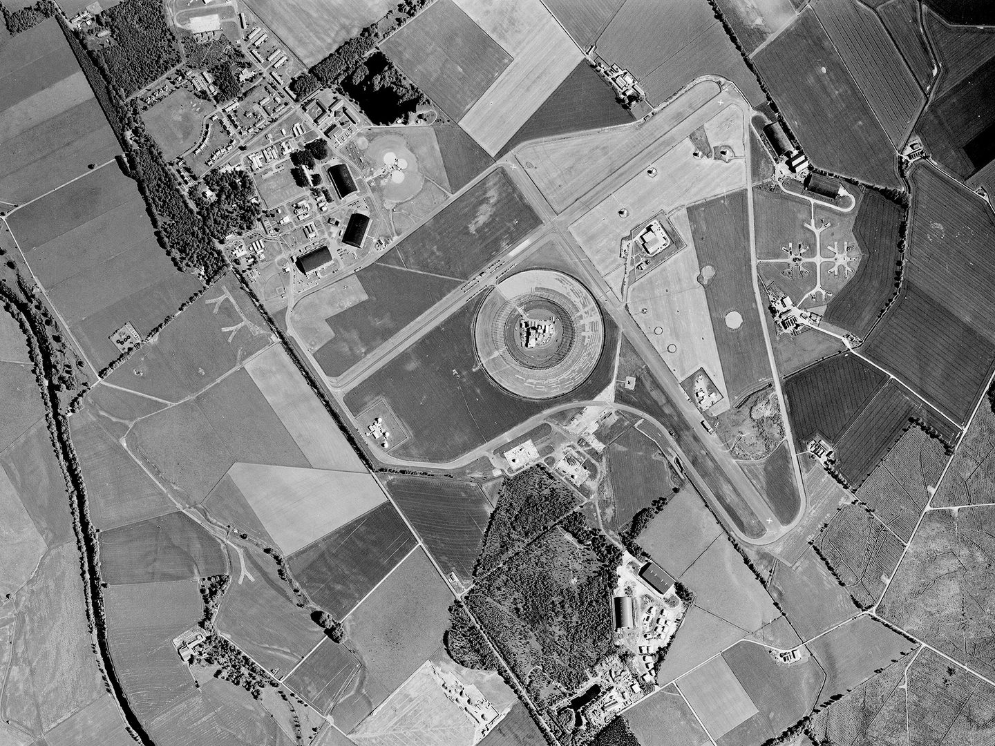 A historic black and white vertical aerial photo of an airfield. 2 runways cross in an X shape in the middle of the image. In the angle of the cross is a circular enclosure, and 3 hangars are visible to the top left.