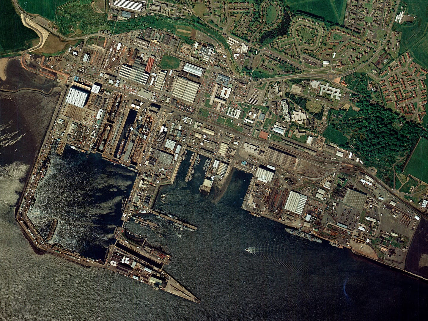A colour aerial photo of a port. Piers jut out into the water, forming a large square enclosure. Numerous ships of various size are docked around the port, including 2 in a dry dock. Large buildings and houses fill the shore.