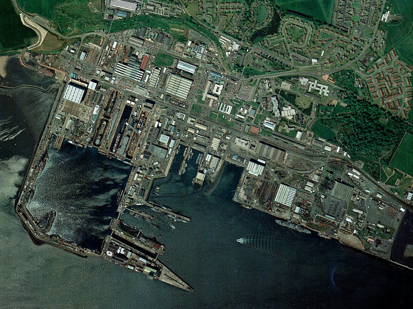 A colour aerial photo of a port. Piers jut out into the water, forming a large square enclosure. Numerous ships of various size are docked around the port, including 2 in a dry dock. Large buildings and houses fill the shore.