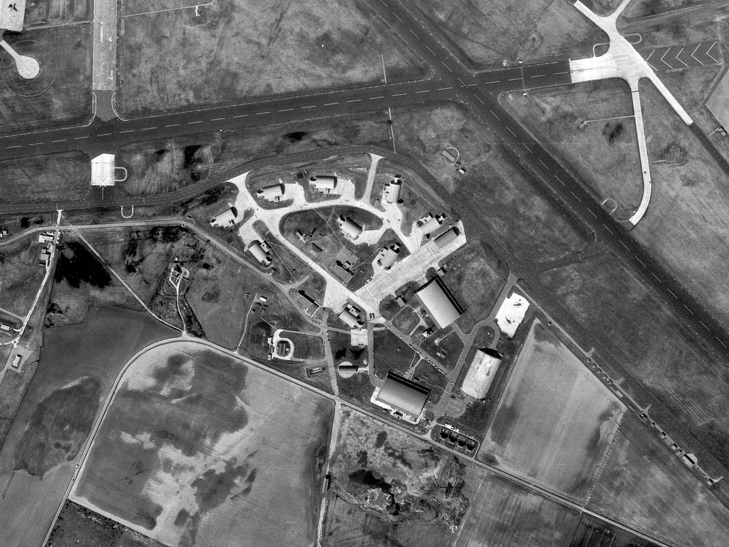 A black and white aerial photo of an airfield. Runways run along the top and right-hand edges. The photo is focussed on a collection of buildings, including 3 large aircraft hangars and 9 smaller, reinforced hangars.